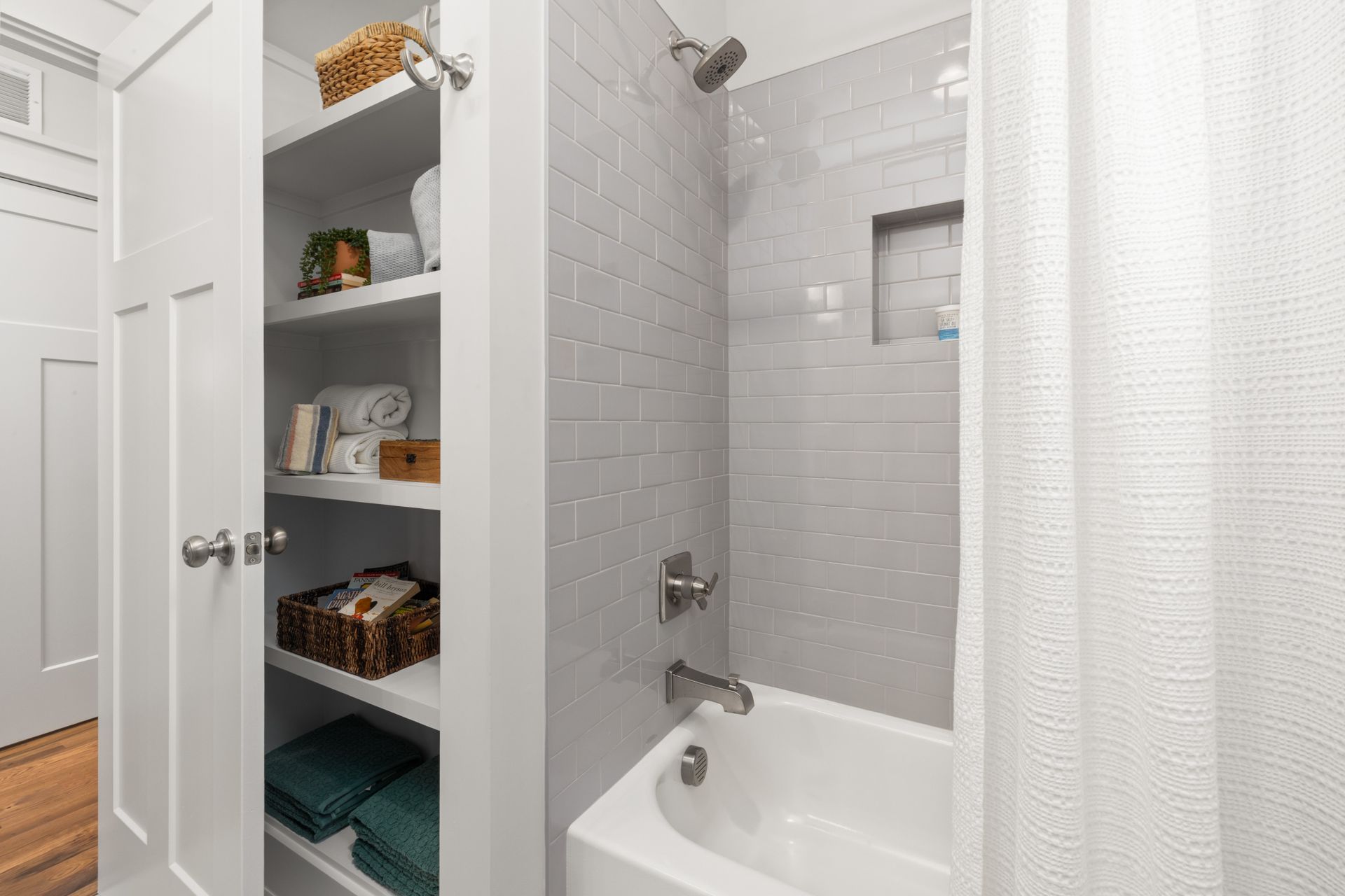 Built-in bathroom linen closet beside a tub with its paneled door open on deep white shelves of rolled and folded towels and woven baskets