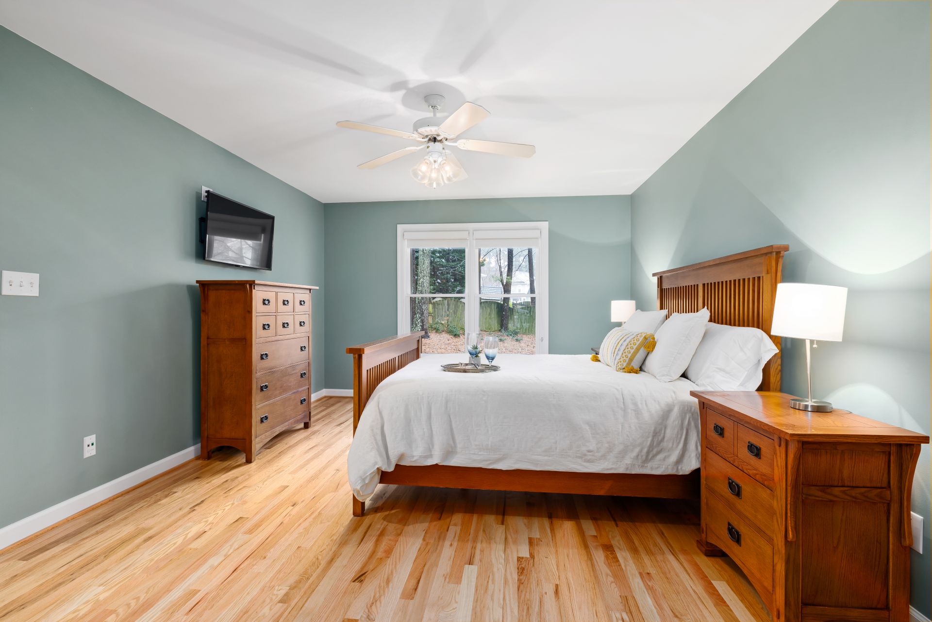 Bedroom with sage-green walls, a pair of white double-hung windows, a ceiling fan with a light kit and a duplex outlet near the floor