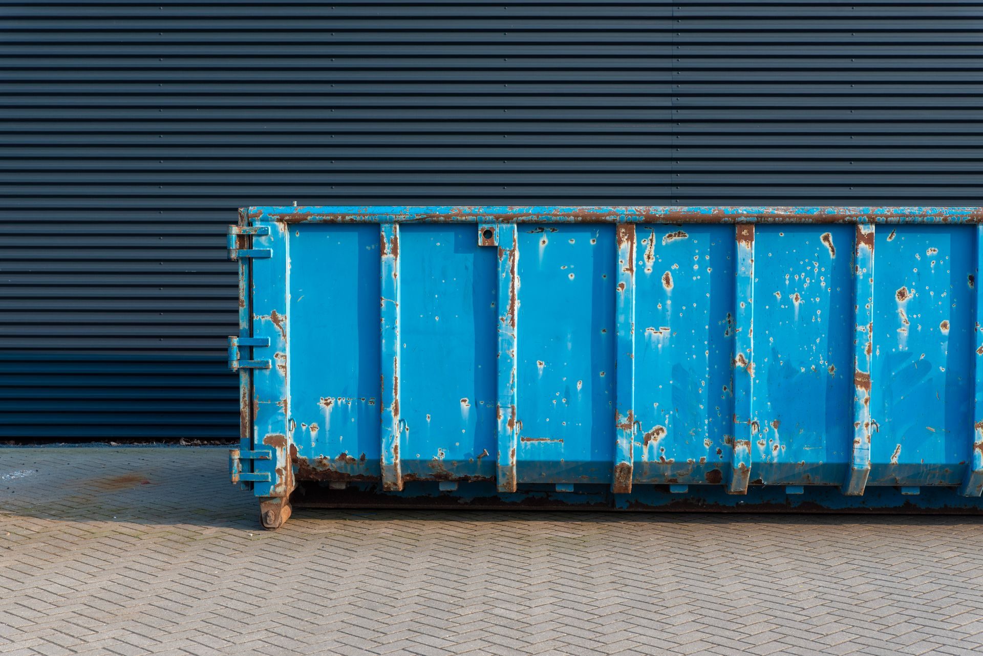 A weathered blue roll-off dumpster with rust spots on gray pavers against a dark corrugated metal wall