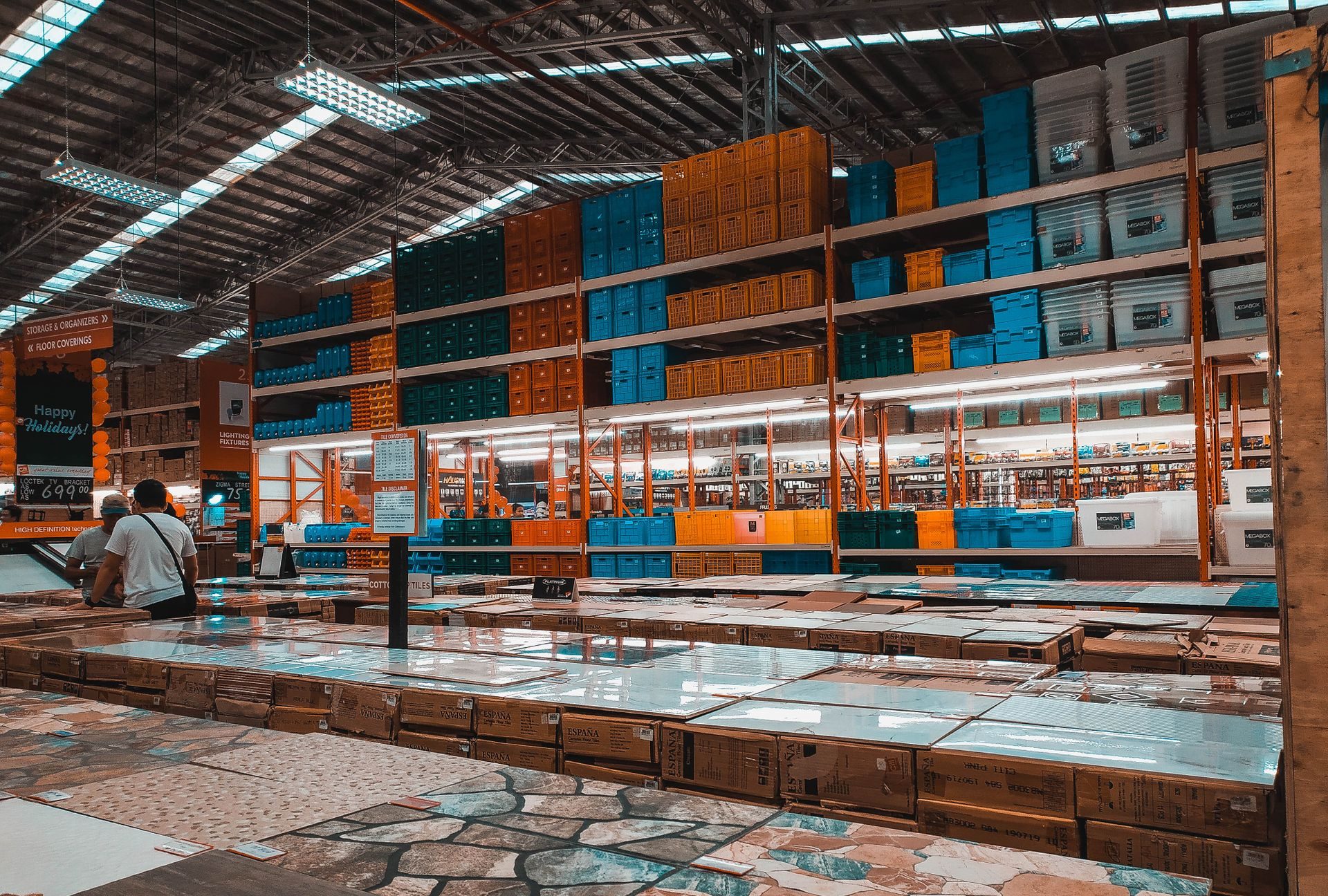 Boxed tile stacked waist-high on pallets on a big-box store floor, with loose sample tiles laid on top of the stacks and tall racks of storage bins behind