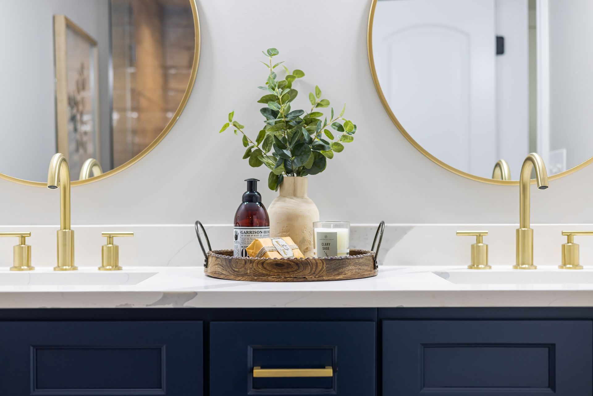 Close view of two brushed-brass widespread faucets, each a separate spout between two handles, on a white quartz top over navy cabinets, under round brass-framed mirrors