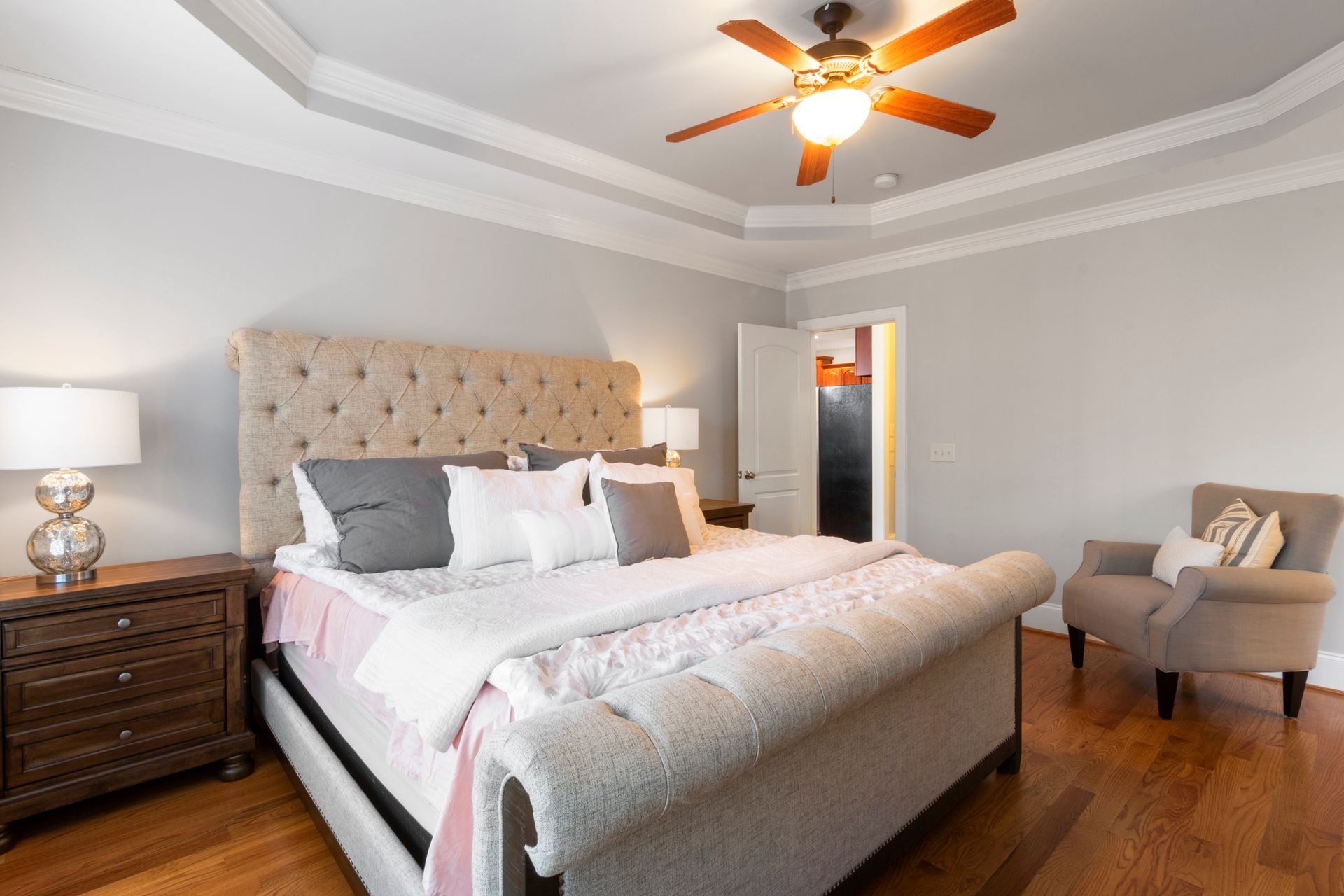 Oil-rubbed bronze five-blade ceiling fan with cherry-finish blades and a glowing frosted glass bowl light, centered in a bedroom tray ceiling above an upholstered bed