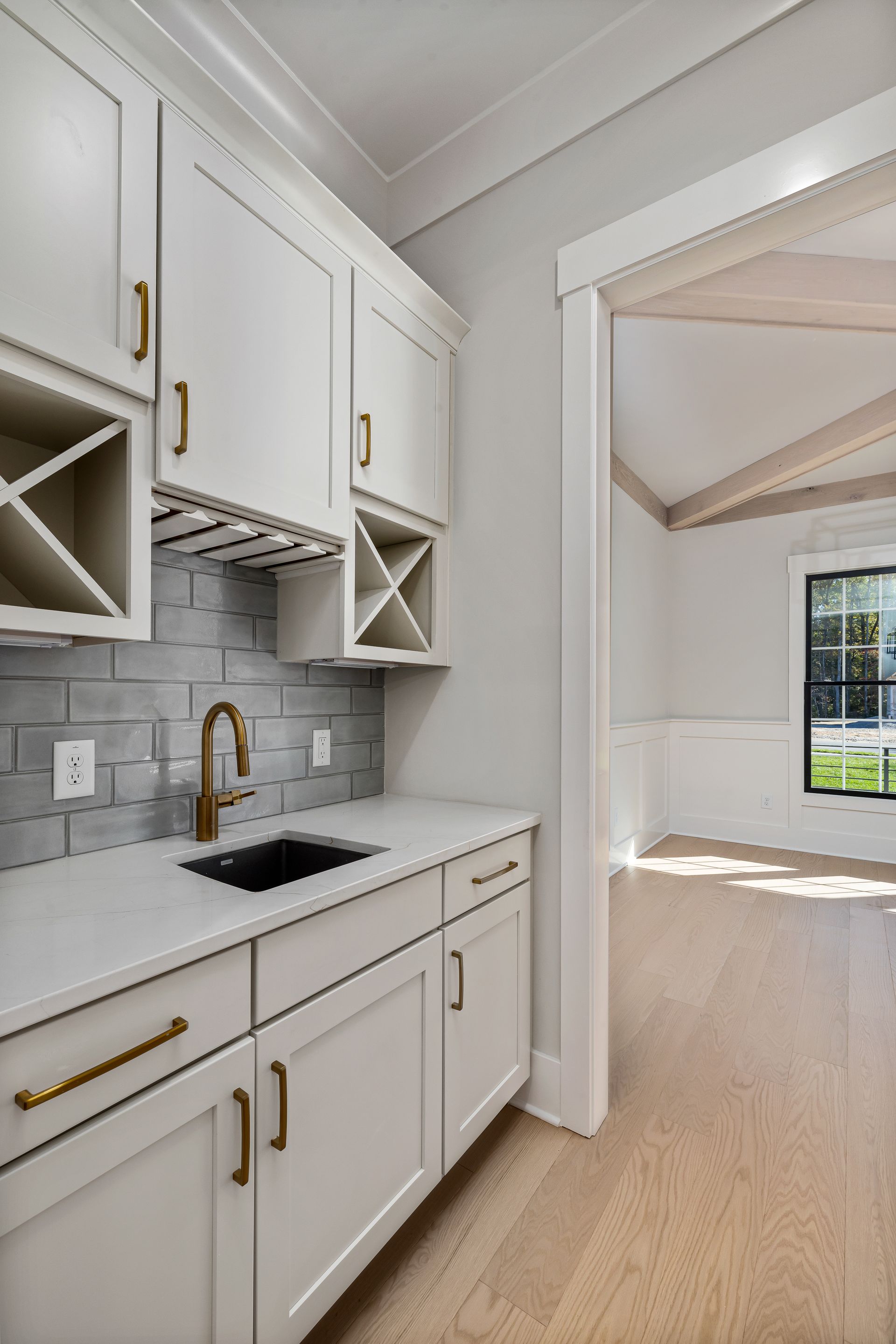Butler's pantry with white shaker cabinets, wine cubbies, a stemware rack, a quartz counter and a black bar sink, opening to a dining room