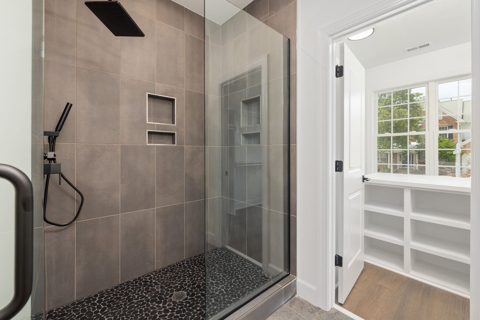 A walk-in shower with large charcoal porcelain wall tile, two recessed niches, a black pebble-mosaic floor with a center drain and a frameless glass door