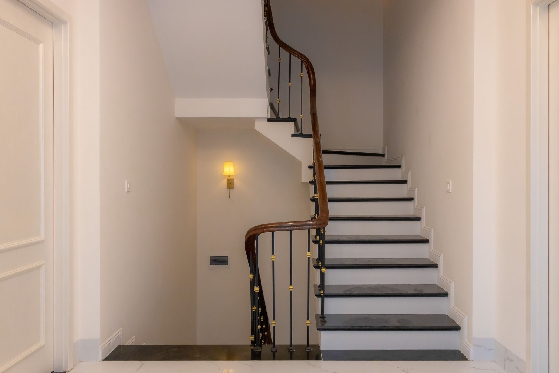 A stair with dark treads and white risers, a curved stained wood handrail with a scrolled starting end, and black iron balusters with gold collars set into the treads