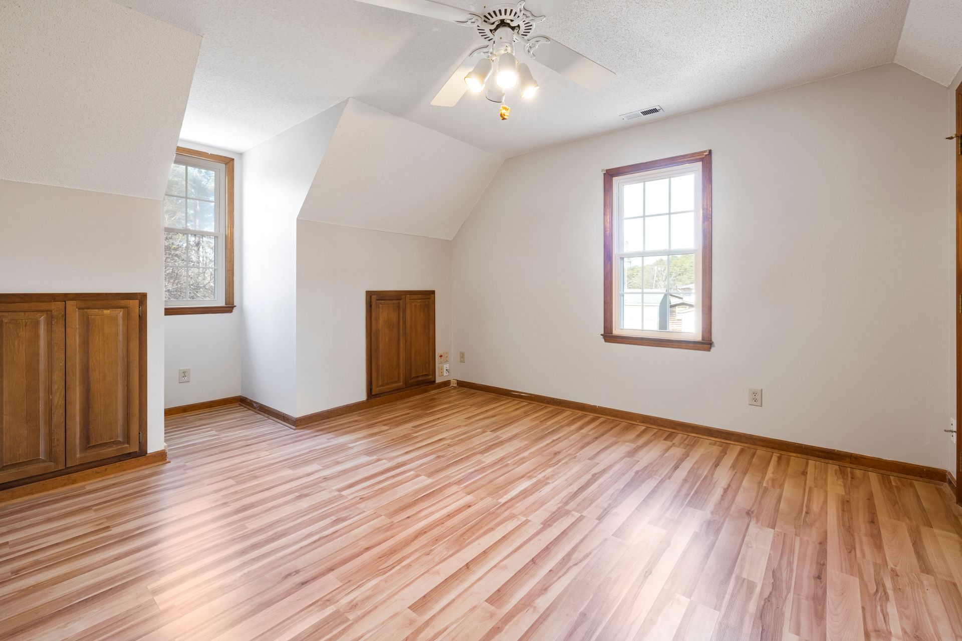 A large empty upstairs bedroom with light plank flooring, a dormer window on the left wall, a second window on the back wall, two wood knee-wall cabinet doors and a ceiling fan with lights