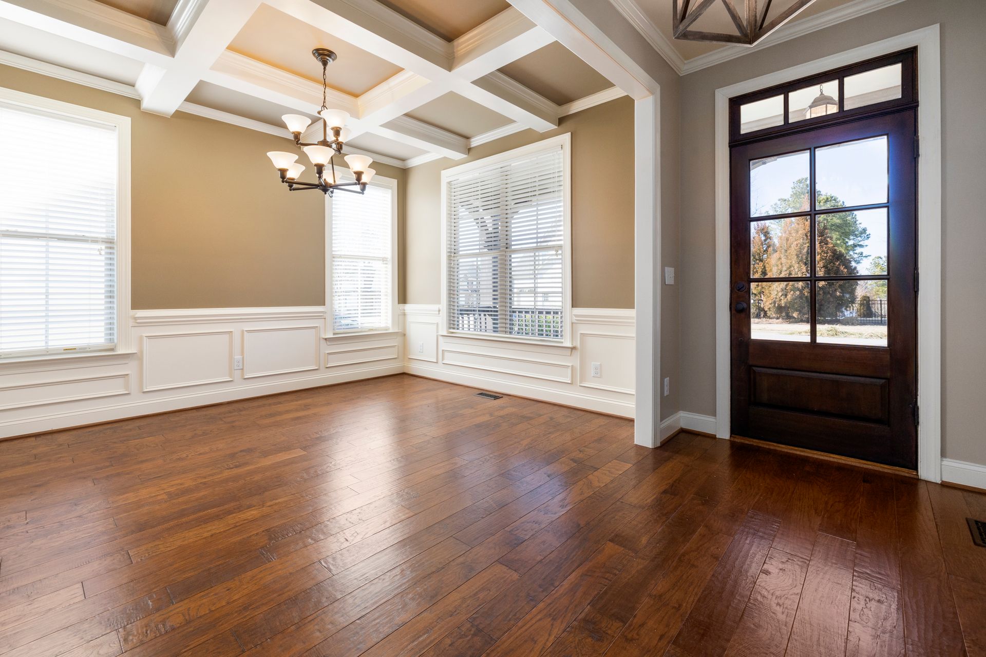 An empty front entry and dining room with taupe walls, white raised-panel wainscoting, a white coffered ceiling, a bronze chandelier, a wood front door with glass lites and hand-scraped hardwood floors