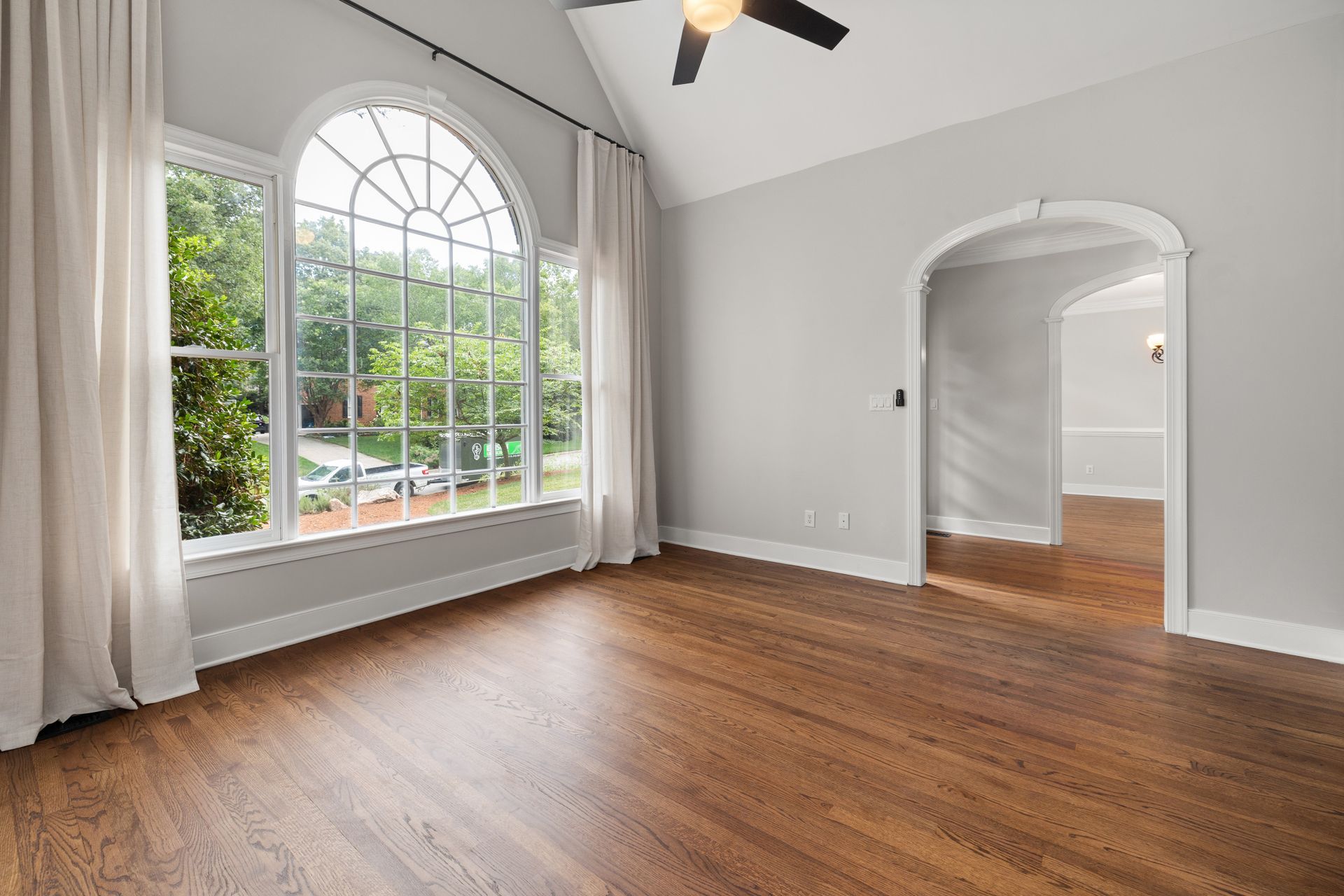 An empty room ready for move-in with a vaulted ceiling, black ceiling fan, arched-top grid window, light gray walls and freshly refinished oak floors