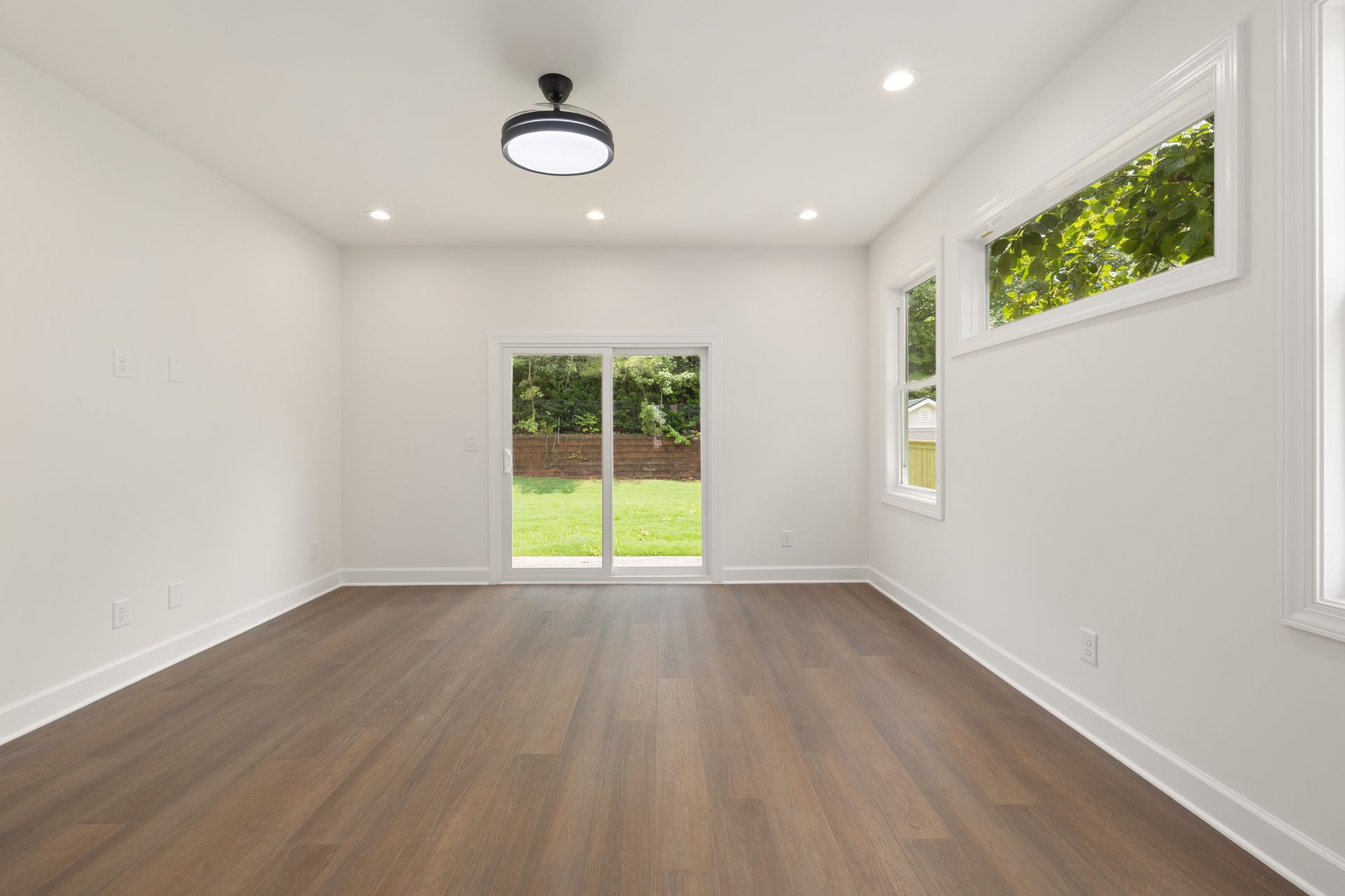 An empty, freshly finished room with dark wood-look vinyl plank flooring, recessed lights, a ceiling fan, a white vinyl sliding patio door, a double-hung window and a long transom window