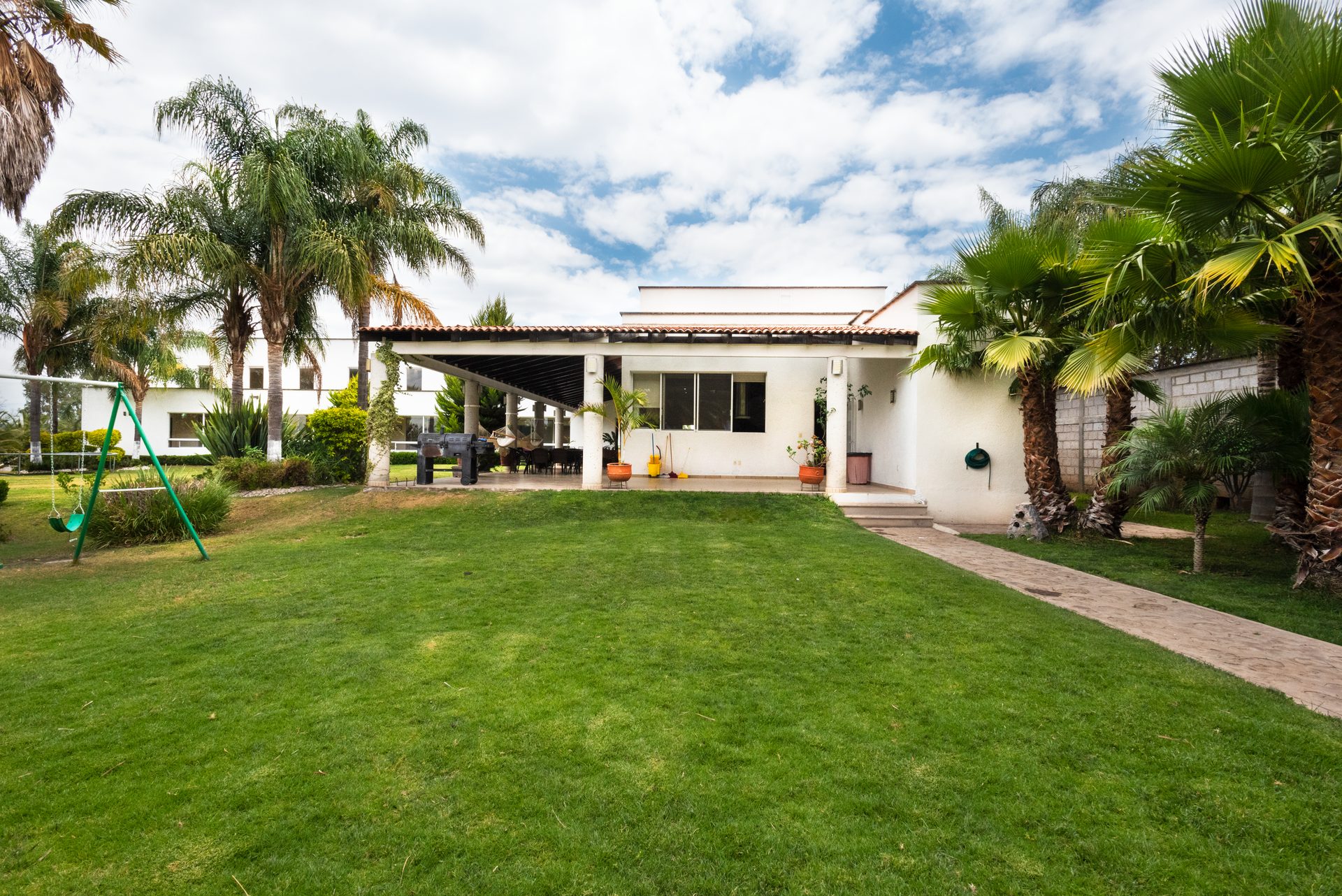 A wide green lawn in front of a single-story white stucco house with a flat roof and a covered patio, framed by tall palms, with a block wall and a paver walk at the right