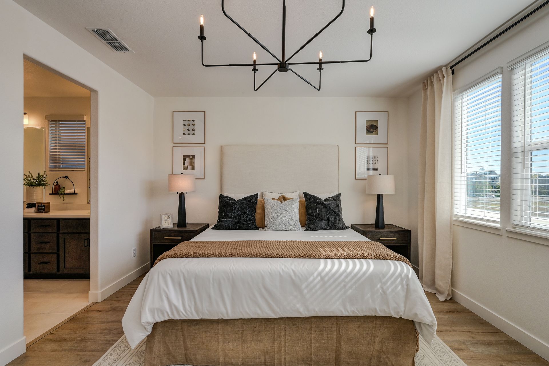 A bright guest bedroom with an upholstered bed, dark wood nightstands, linen drapes, wood-look plank flooring and an open doorway into an ensuite bath