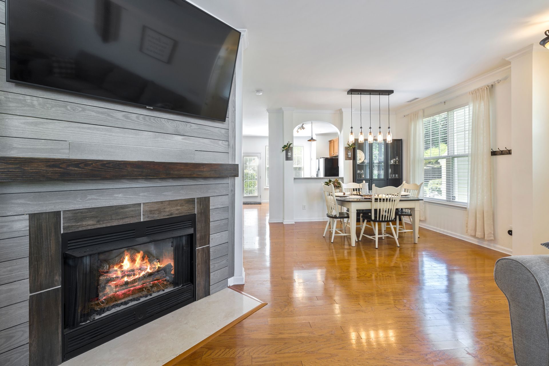 A living room with a gray shiplap fireplace wall and TV opening across glossy oak floors to a dining table, with a half-height wall, a flat ledge cap and a square column framing an arched pass-through to the kitchen