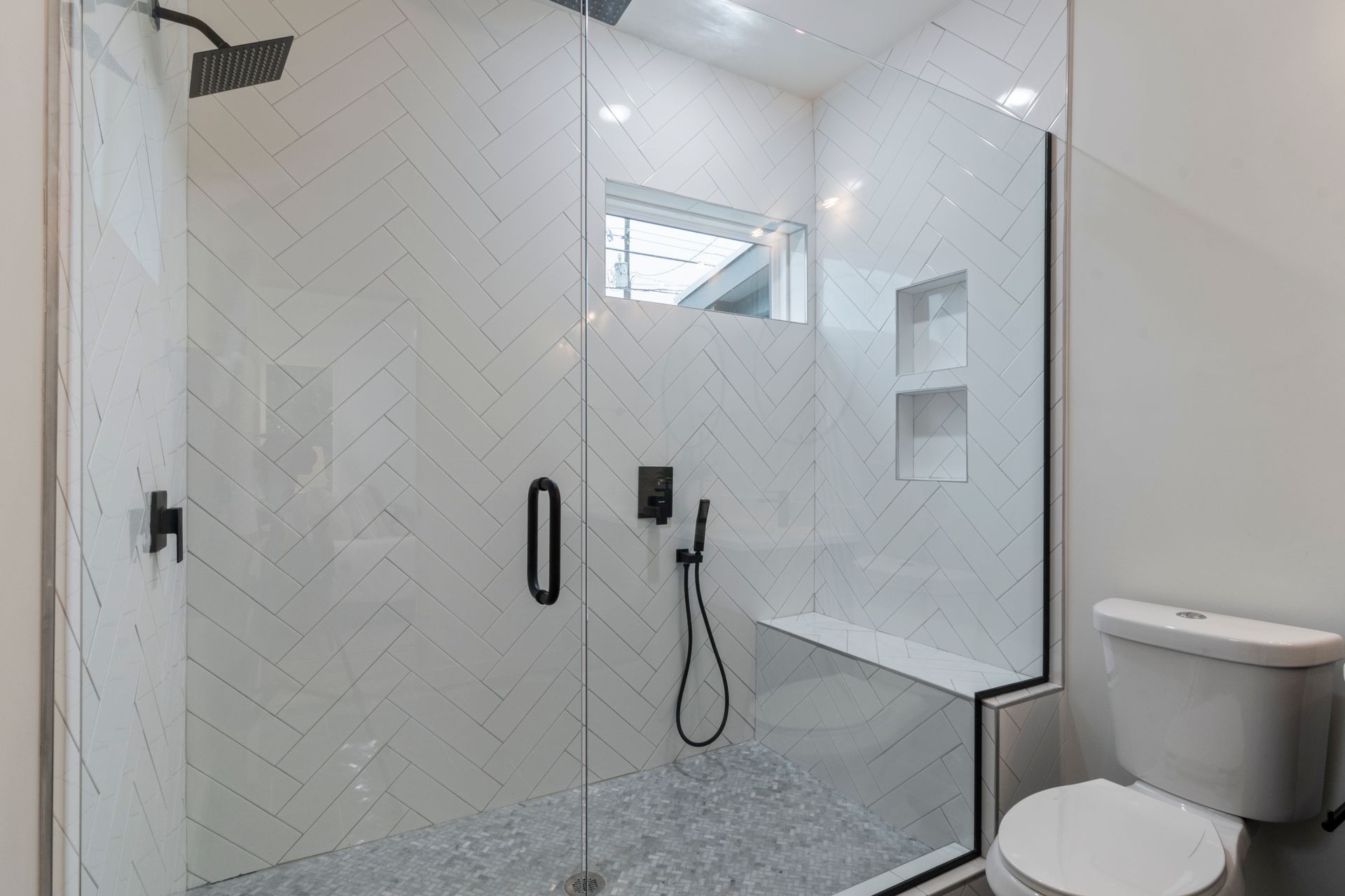Walk-in shower with a frameless glass door and fixed panel, white herringbone wall tile, a gray marble herringbone mosaic floor with a center drain, a corner bench and two recessed niches