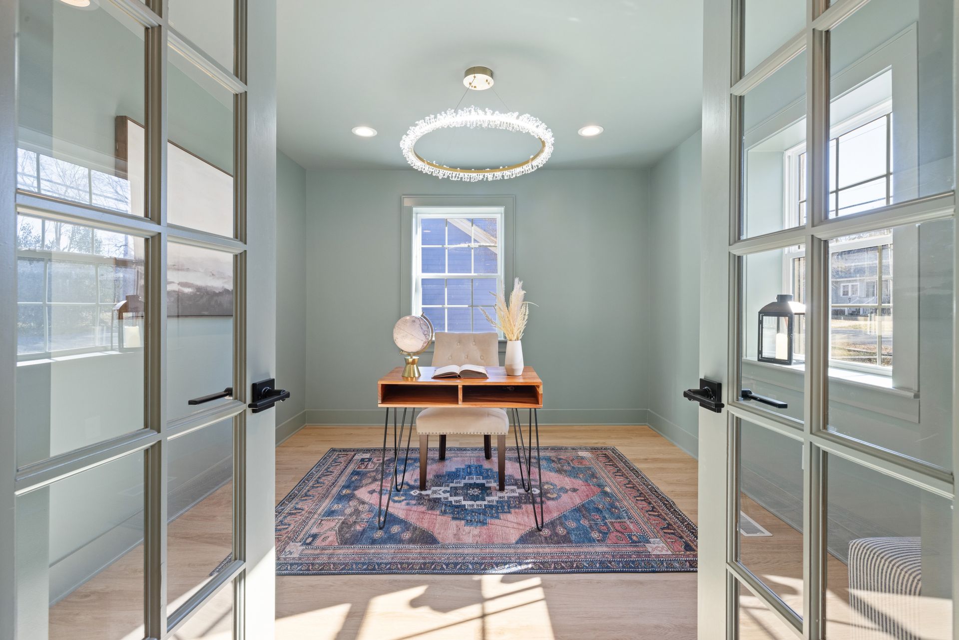 Small home office seen through a pair of open glass-panel French doors, with a wood desk, a chandelier and a floor supply register