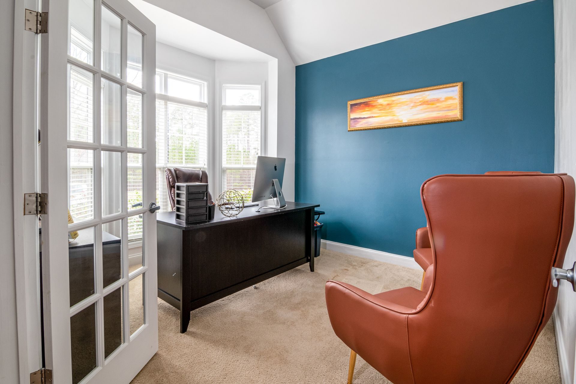 A home office seen through a pair of open white French doors with divided glass panes, with a teal accent wall, a dark wood desk with a computer monitor, a tan leather lounge chair, beige carpet and a white bay window on the left wall