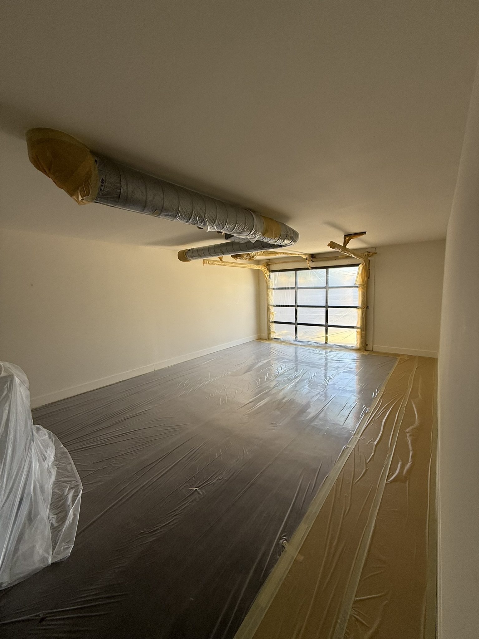 Interior room prepped for painting with plastic sheeting run wall to wall across the floor and exposed ductwork wrapped in paper and tape