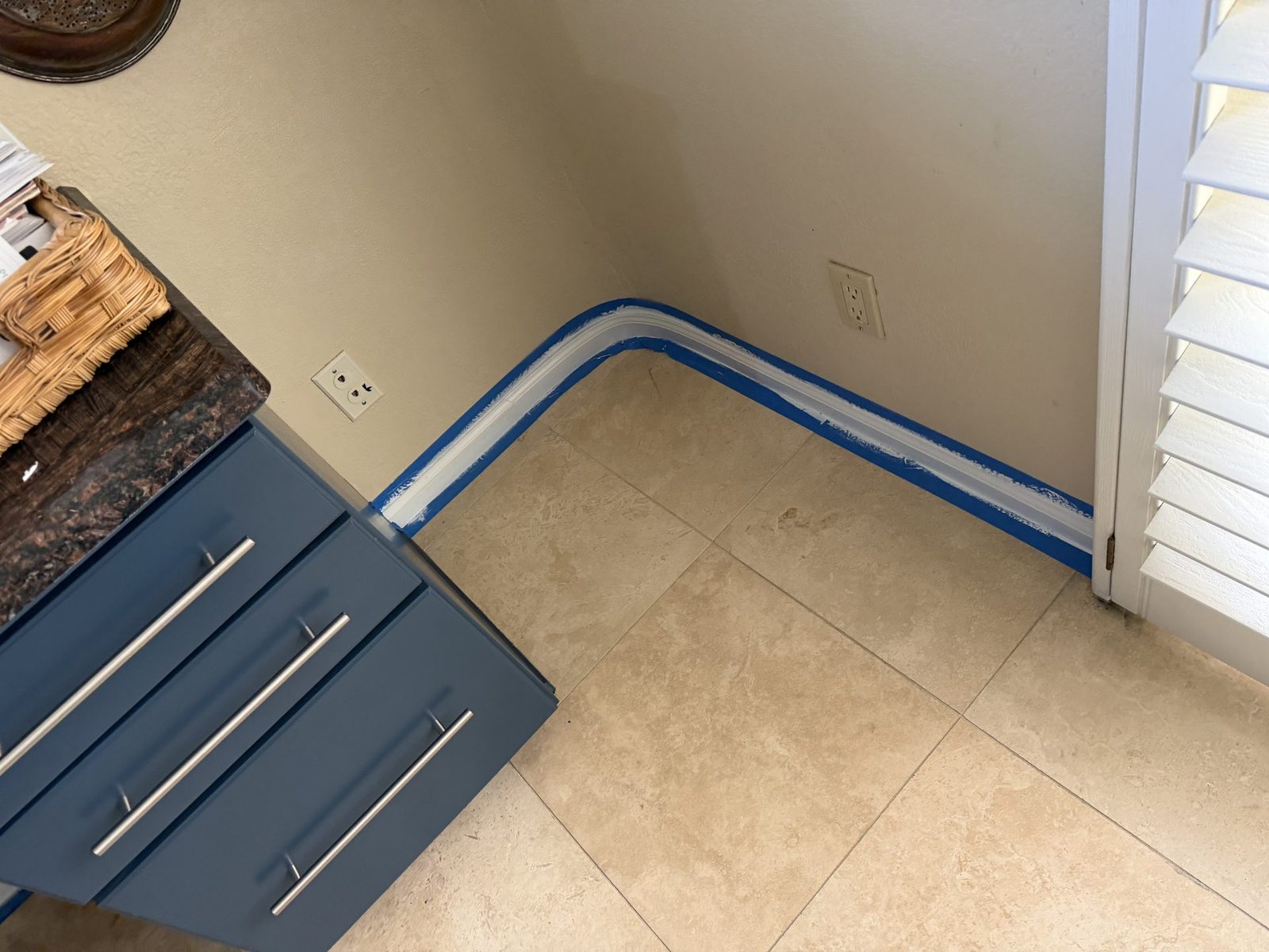 Blue painter's tape pressed onto travertine tile along a freshly painted baseboard next to a navy built-in desk