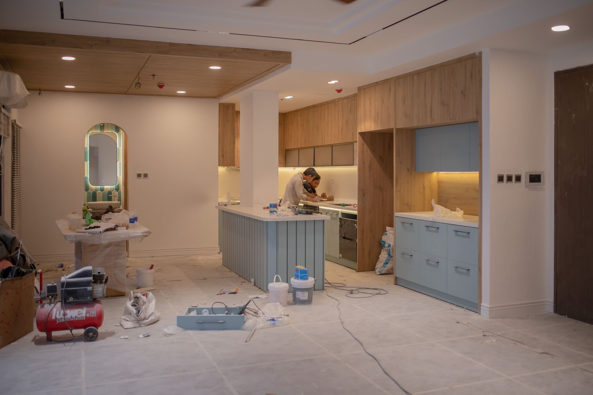 A kitchen mid-install with flat-panel wood-veneer upper cabinets, pale blue base cabinets and island, a paper-covered floor, and an air compressor and tool cases in the foreground