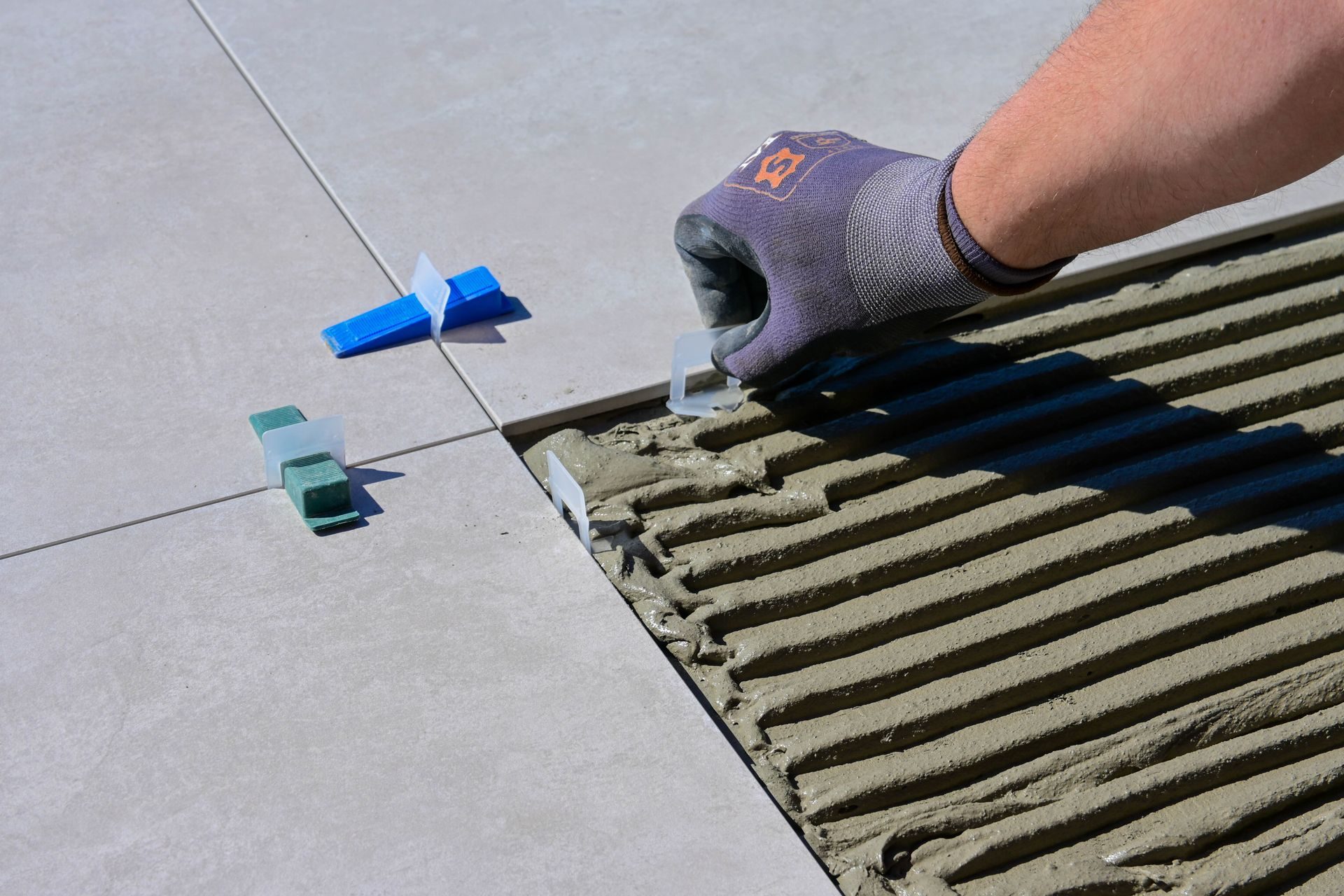 A gloved hand setting a large-format light gray porcelain tile into combed gray thinset, with blue wedge leveling clips and green caps in the joints of the tiles already laid