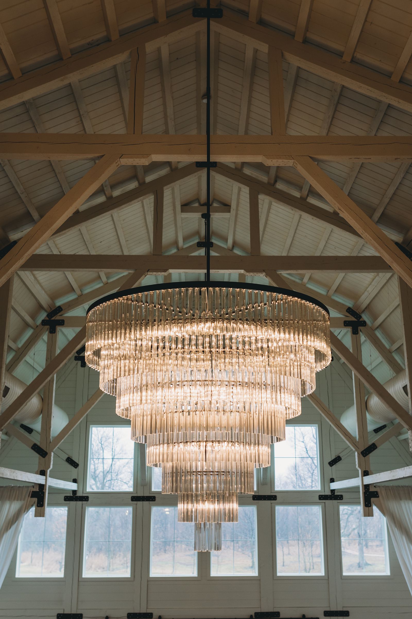 Large multi-tier chandelier of clear glass rods on a black ring frame, hung on one long black rod from the ridge of a cathedral ceiling with whitewashed timber trusses and tall gable windows