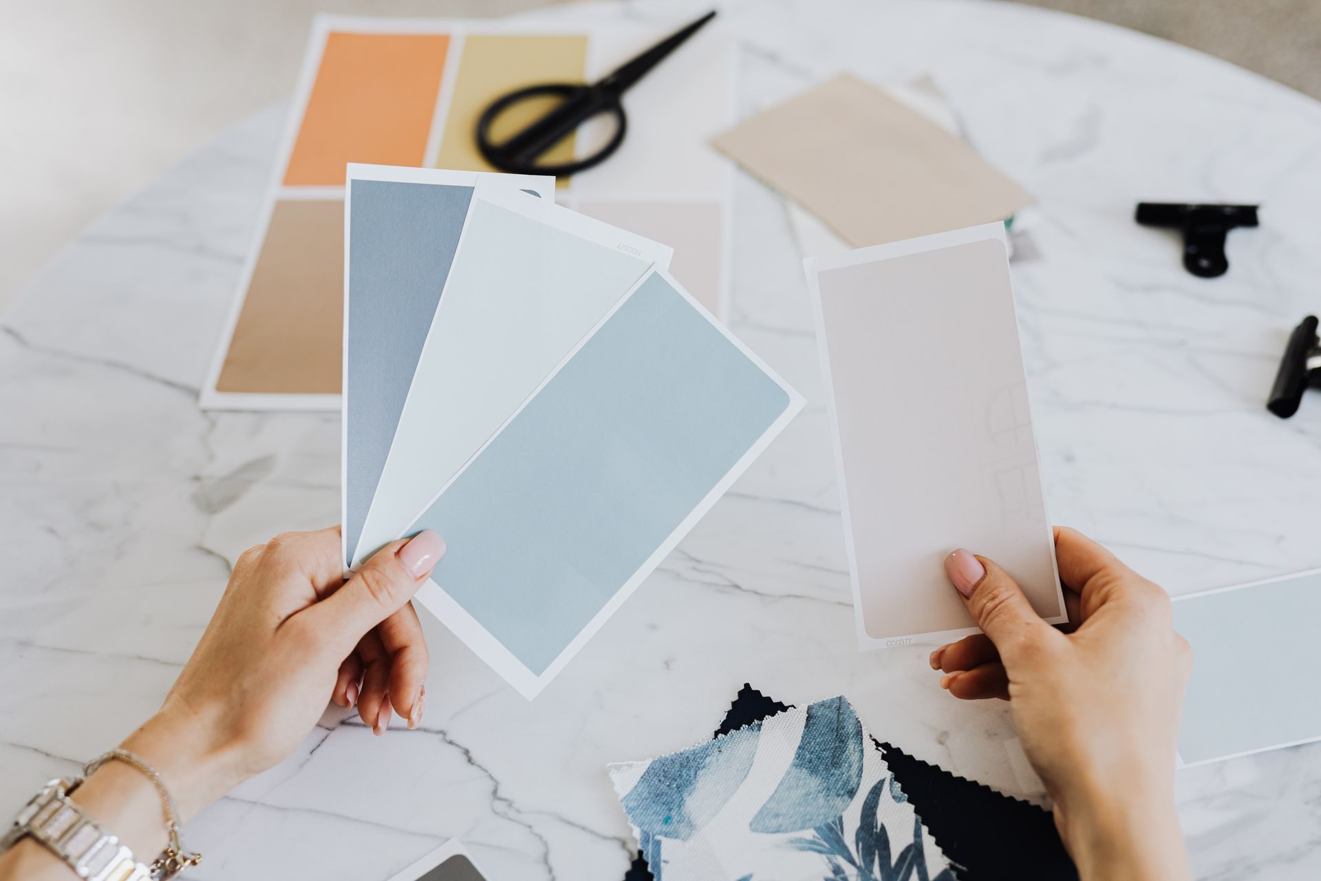 Hands fanning out blue-gray, white and blush paint swatch cards over a white marble table, with more swatches, scissors, binder clips and a fabric sample nearby