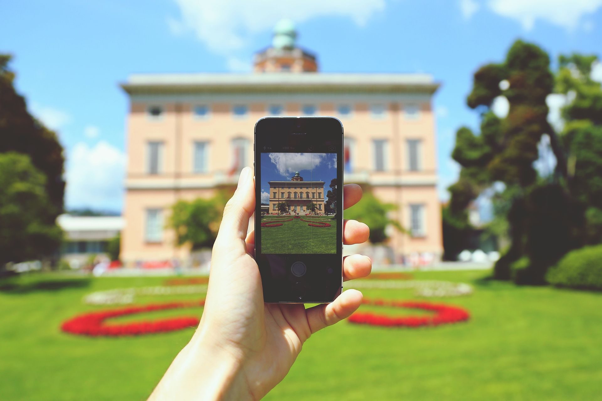 A hand holding up a phone whose screen shows a mown green lawn with curving red flower beds in front of a peach-colored house with a domed tower
