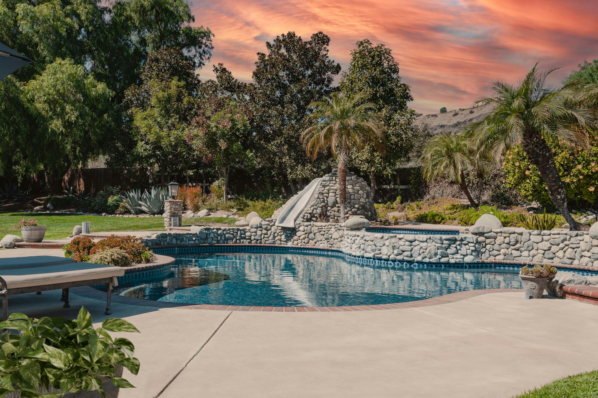 Light concrete pool deck curving around a freeform pool with a river rock wall, slide, palms and agave
