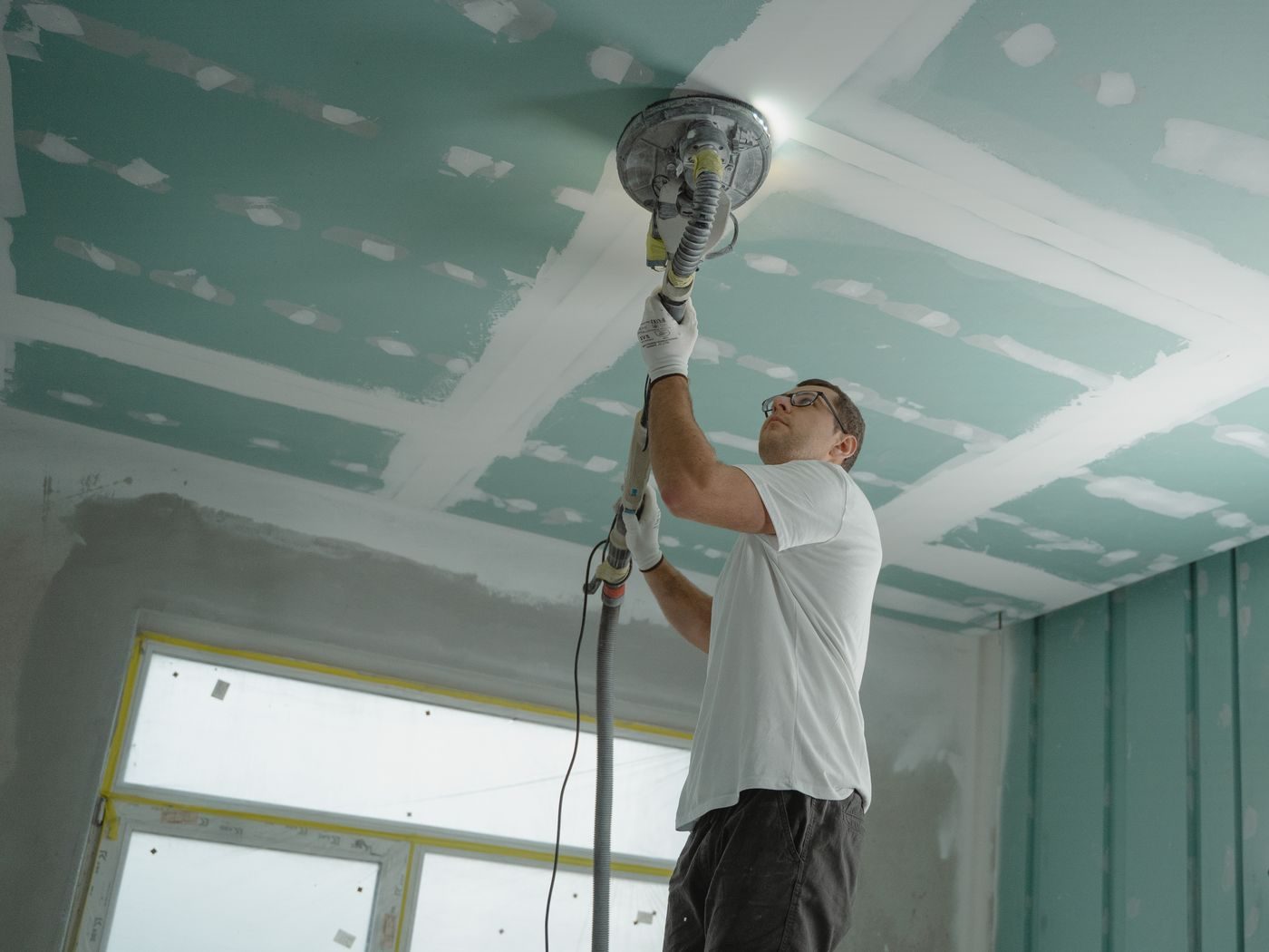 Worker sanding a taped and coated ceiling overhead with a pole sander and a work light raking across the surface