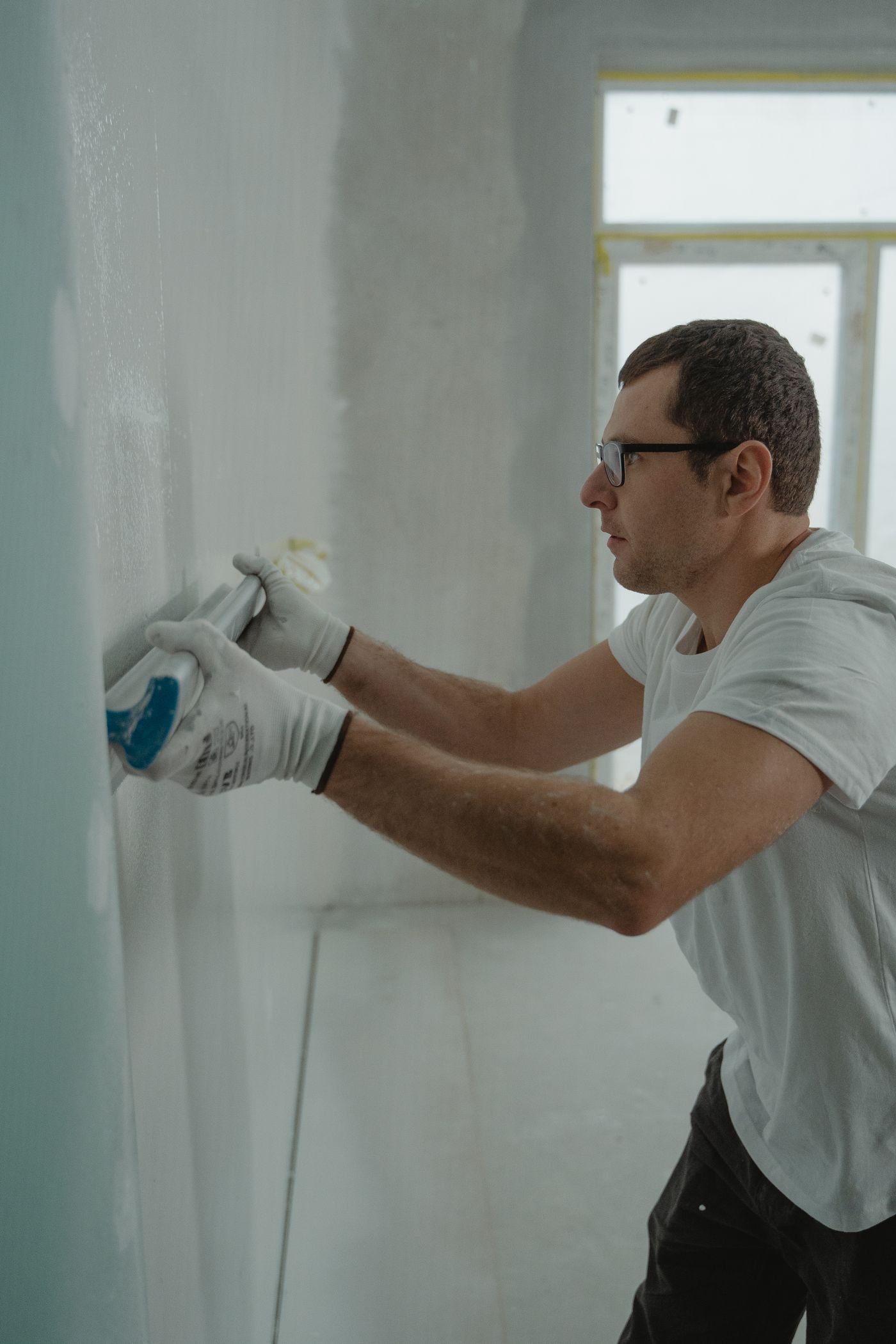 Worker in gloves feathering joint compound outward across a wall so the repair blends into the surrounding surface