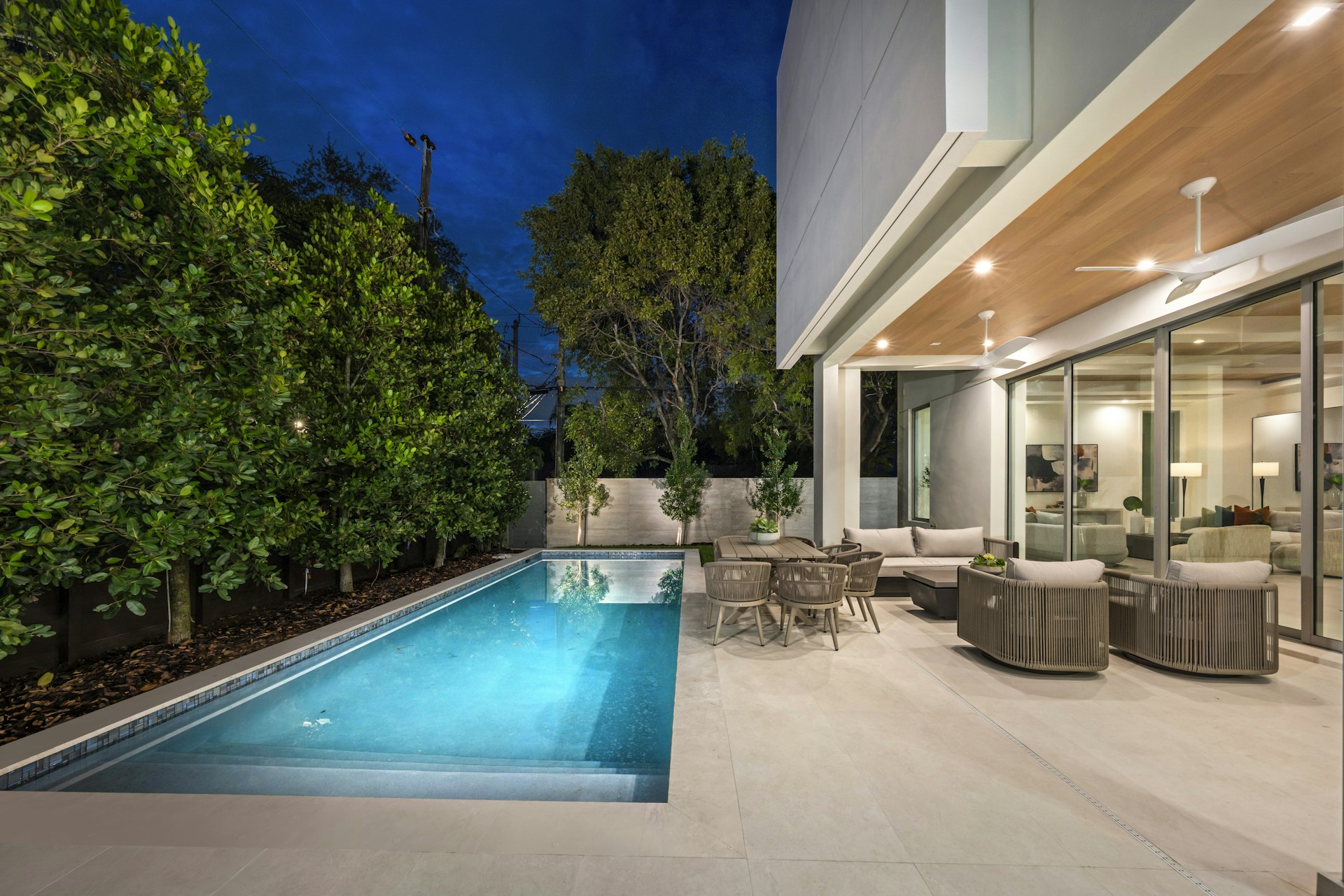 Large-format light gray paver deck along a lap pool with outdoor dining and lounge seating under a wood-soffit patio cover at dusk