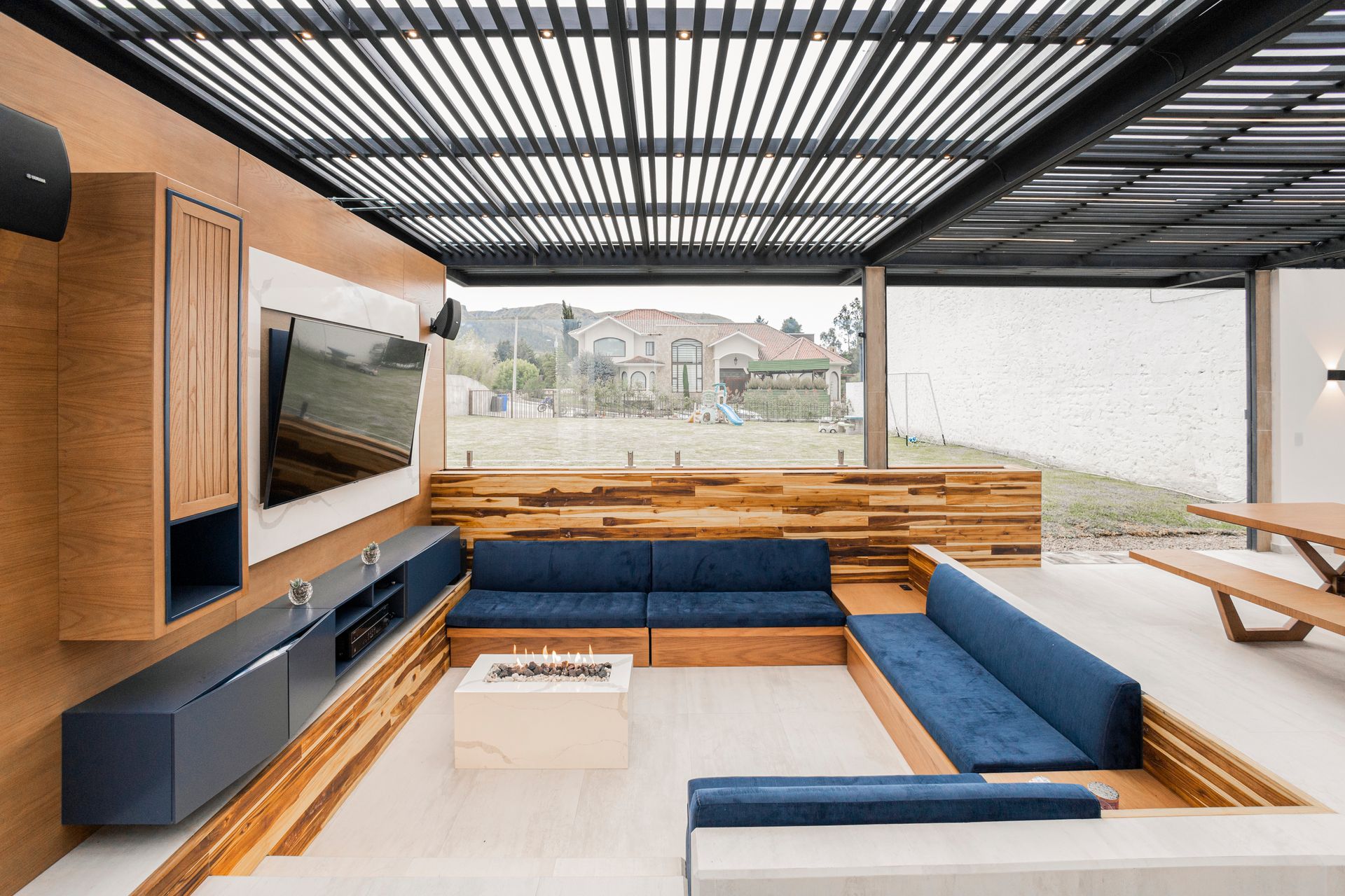 Black aluminum louvered roof over a sunken outdoor lounge with built-in seating, a fire table and a wall-mounted TV