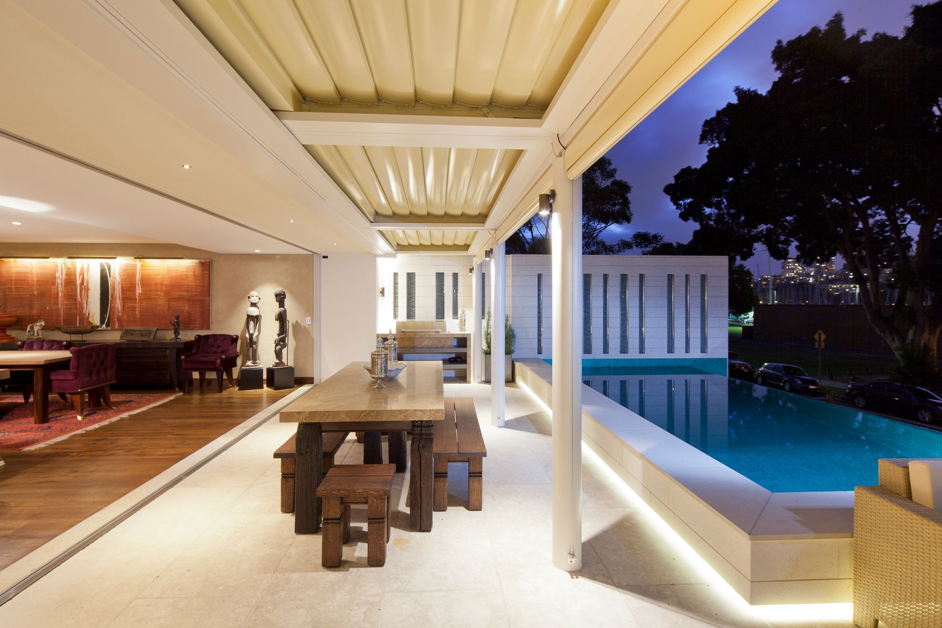 White louvered roof running from the house to the pool edge, with lit dining table and indoor-outdoor living room at dusk