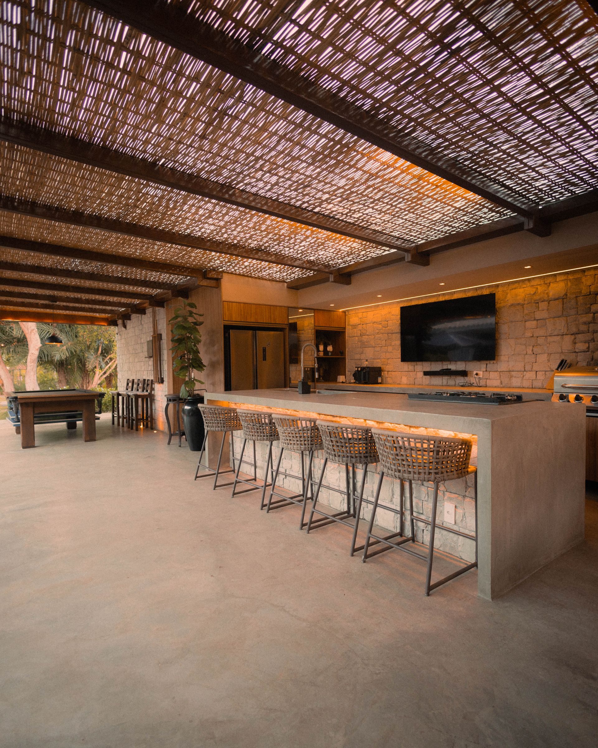 Long outdoor kitchen bar counter with stools, a built-in gas cooktop and a stone veneer base under a shade structure