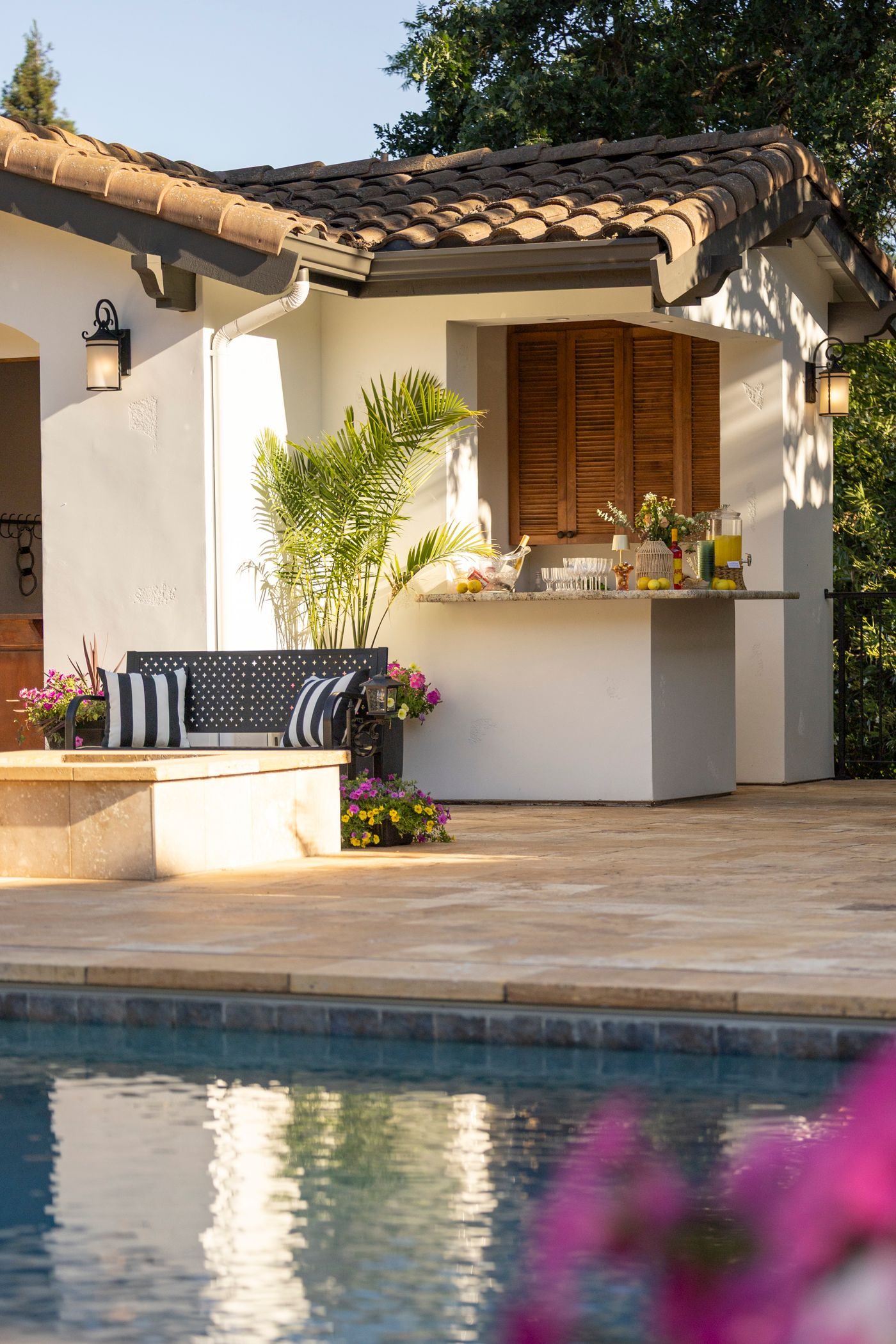 Stuccoed outdoor kitchen island with a bar counter and stools, built beside a pool on a travertine deck under a tile roof overhang