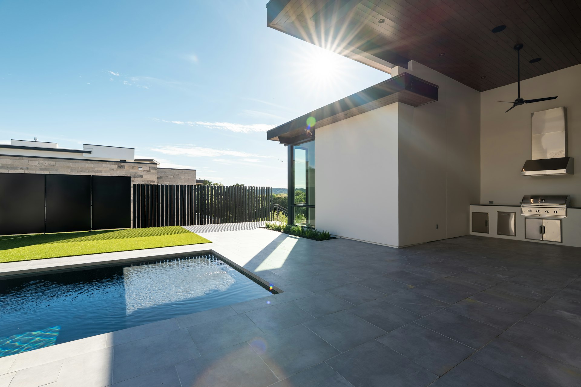 Tall solid patio roof with a wood ceiling and fan over a built-in grill and vent hood, opening onto a pool and artificial turf