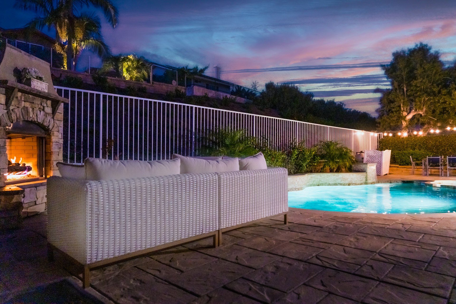 Stamped concrete pool deck in a slate pattern at dusk with a stone fireplace, outdoor sofa and lit pool