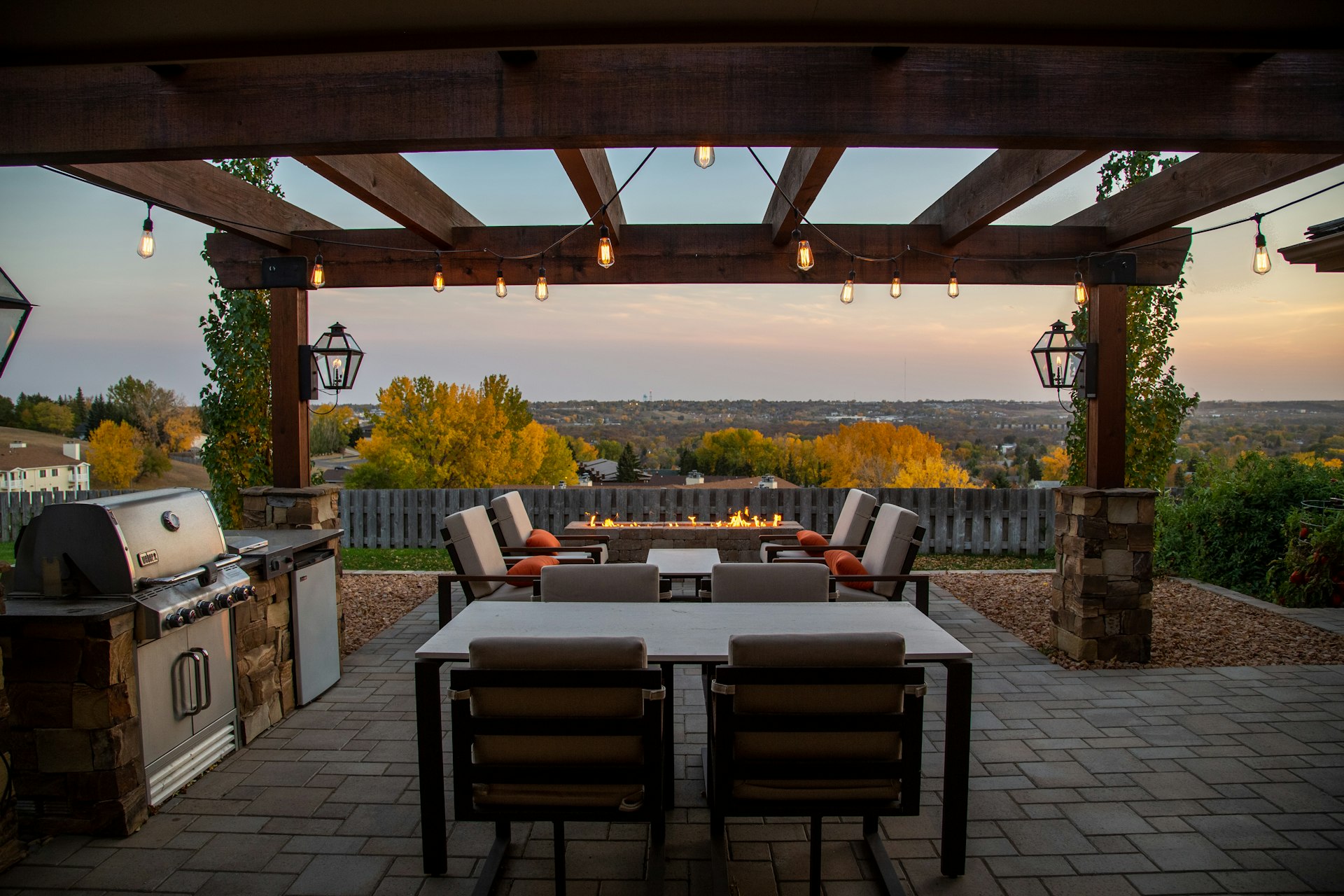 Timber pergola on stone-clad posts over a paver dining patio with a built-in grill and a fire table beyond