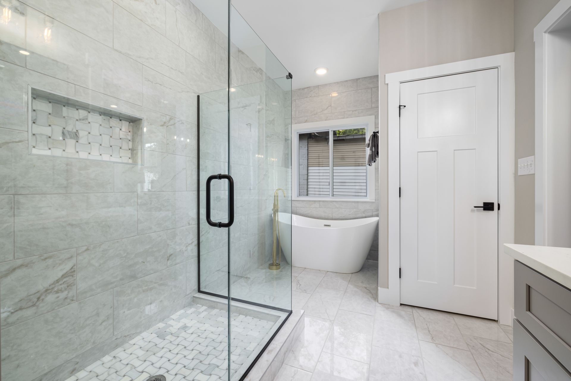 Walk-in shower with large-format gray-veined porcelain walls, a niche lined in basketweave marble mosaic that matches the floor, a low tiled curb and a frameless glass door, with a freestanding soaking tub behind it