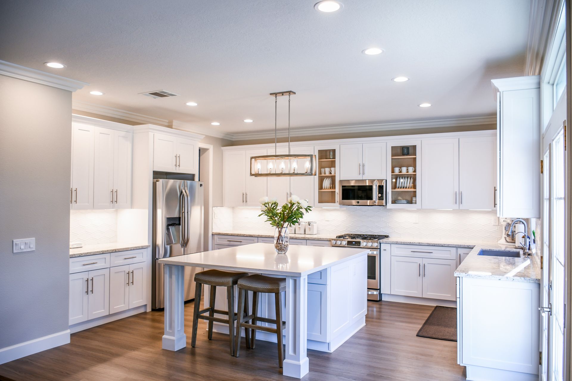 White shaker kitchen with a grid of round recessed ceiling lights, warm under-cabinet lighting on a hexagon tile backsplash and a linear brushed-nickel chandelier over a white quartz island