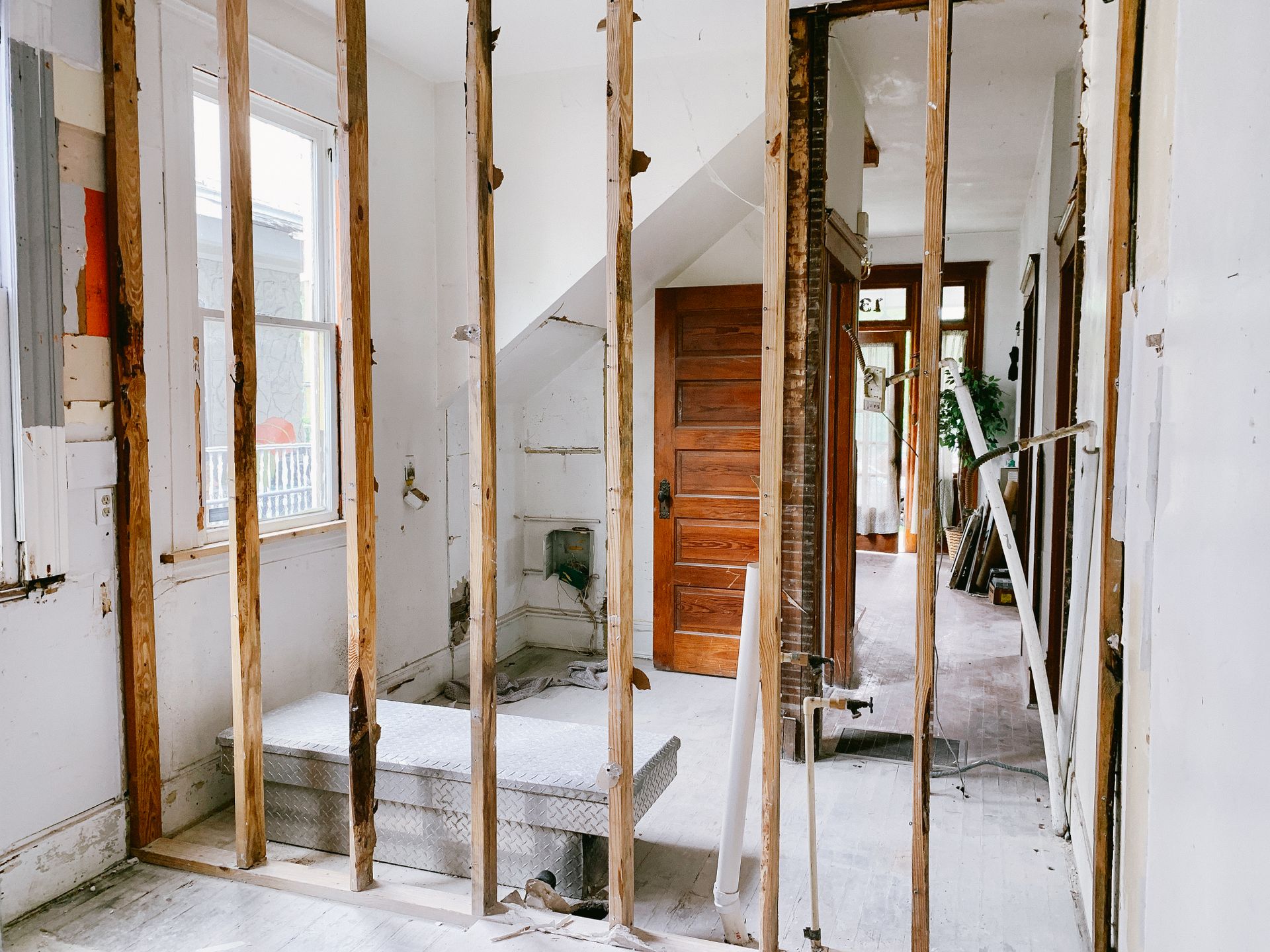 An older house mid-remodel with a wall opened to water-stained studs, capped plumbing stubs and PVC pipe coming up through the floor, an electrical box hanging open and a job box in the hallway