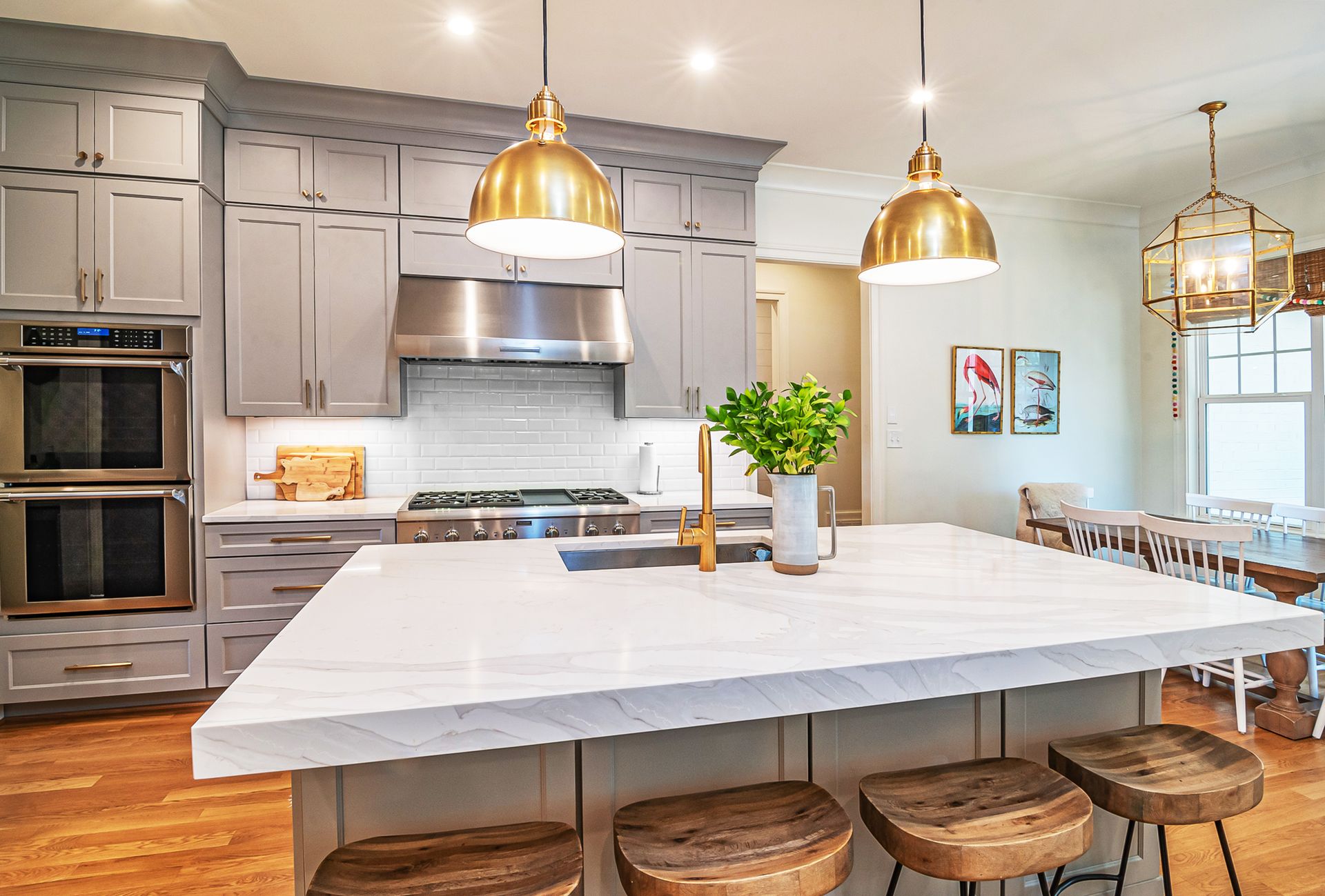 A finished kitchen with gray shaker cabinets to the ceiling, white subway tile backsplash, stainless hood and range, double wall ovens and a white quartz waterfall island under brass dome pendants