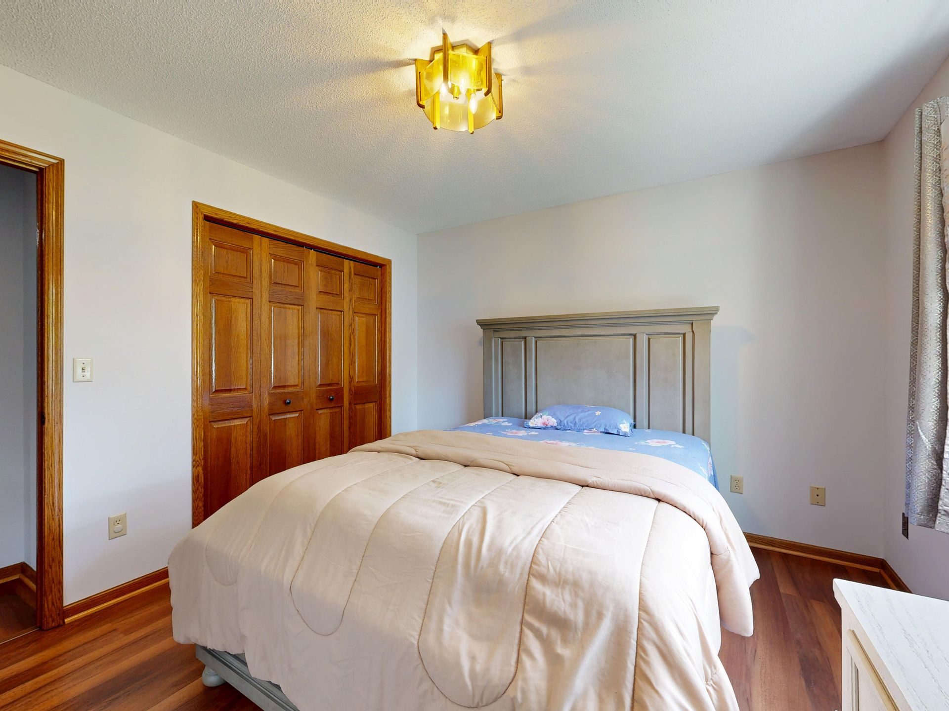 A small bedroom with a bed in a cream comforter, a stained wood bifold door on a reach-in closet on the side wall, an open door to the hall and a wood floor