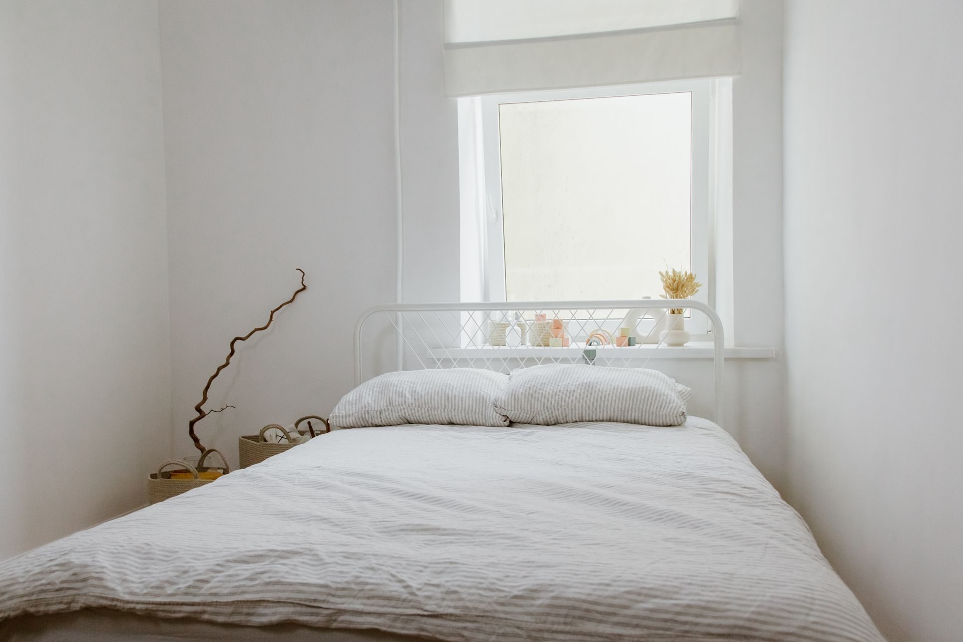 A small white bedroom with a white metal-frame bed under a single window, its roller shade pulled partway down and a deep sill just above the headboard