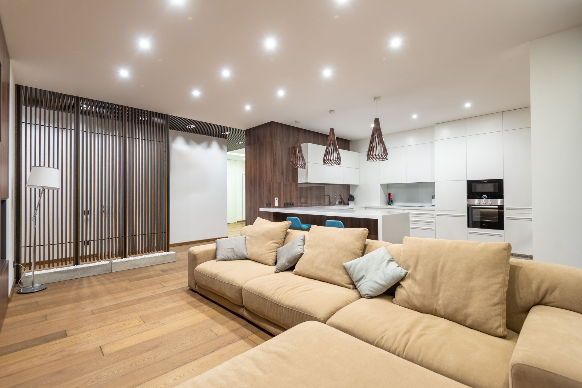 Living area and kitchen with a dozen small square recessed LED downlights in a flat white ceiling and walnut cone pendants over a white quartz peninsula