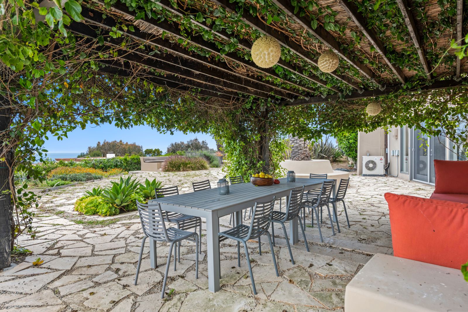 A dark-stained wood pergola cover with closely spaced rafters on a heavy beam over a flagstone dining patio beside a stucco house