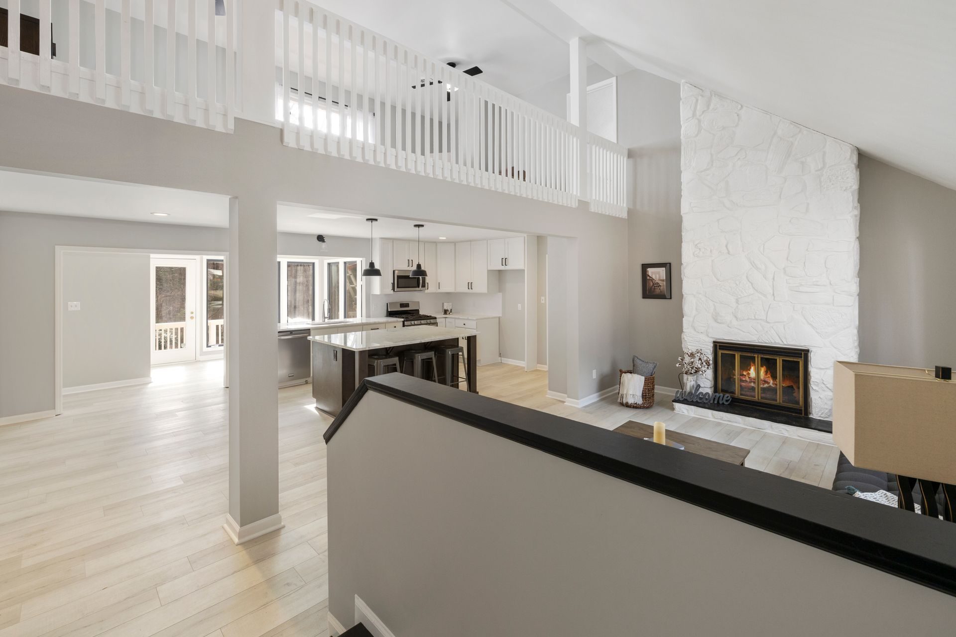 From the top of a stair, a drywall half wall with a dark wood cap runs across a great room with a white kitchen and island, a white-painted stone fireplace and an upstairs loft railed with white balusters