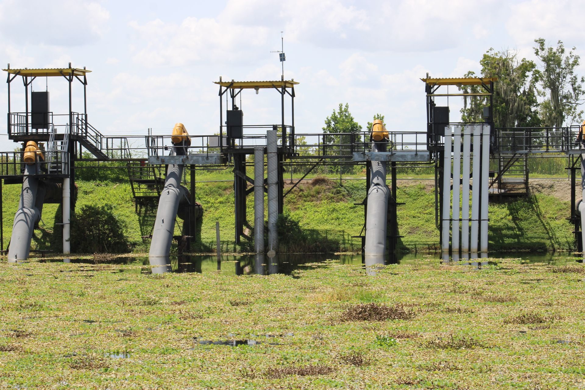 A row of steel water-control structures on a grassy bank, with gray pipes dropping into shallow water, yellow valve housings on top and railed walkways and stairs above