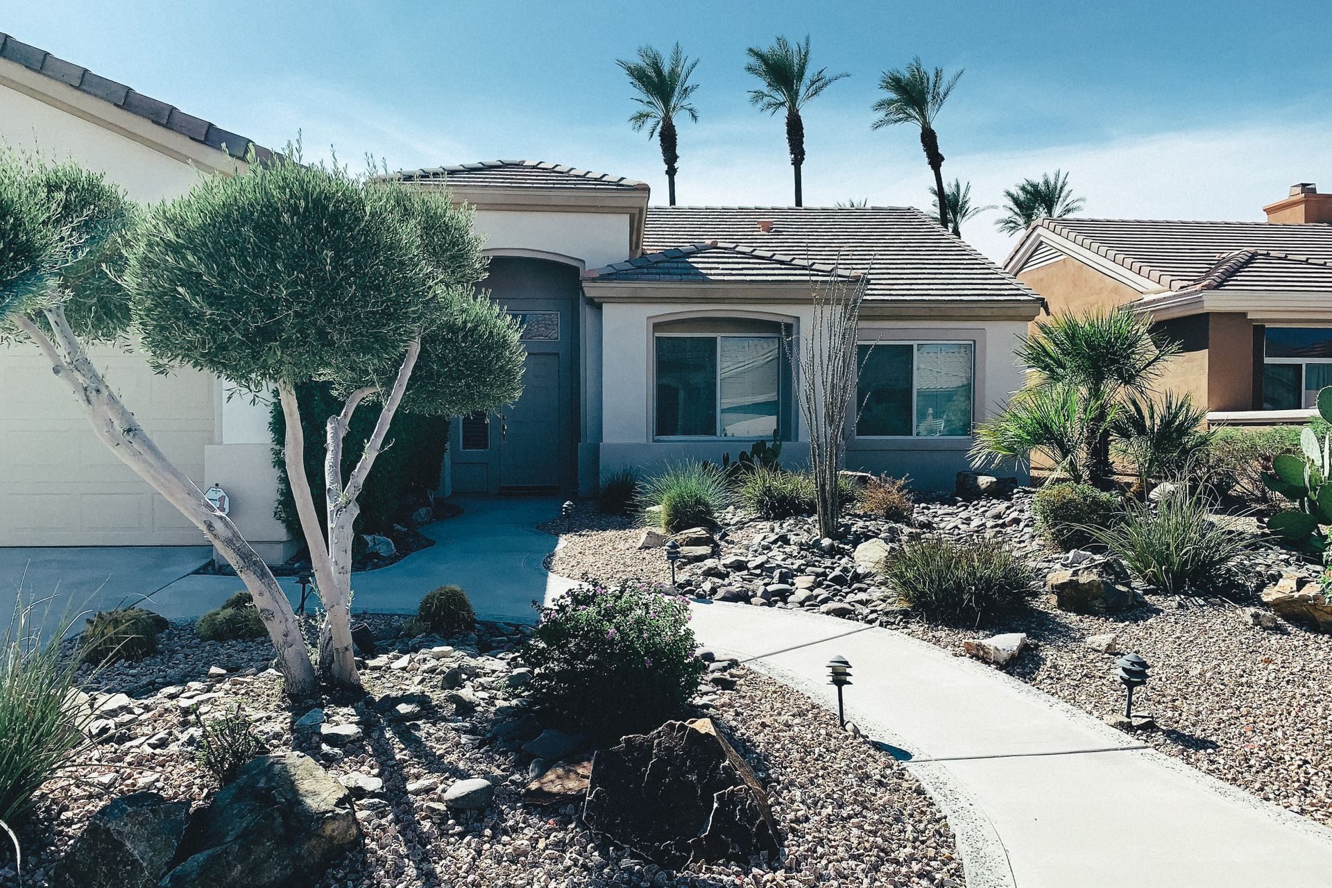 A single-story tan stucco home with a concrete tile roof and an arched entry, behind a gravel-and-boulder desert front yard with olive-style trees, path lights and a curving concrete walk