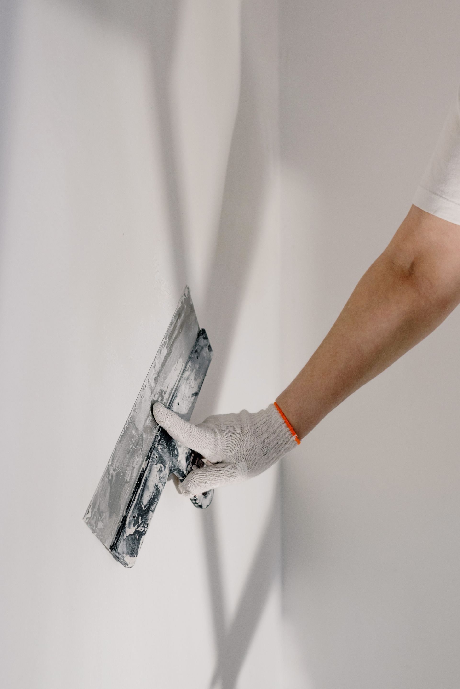 Gloved hand drawing a wide steel taping knife across smooth white drywall to feather a coat of joint compound