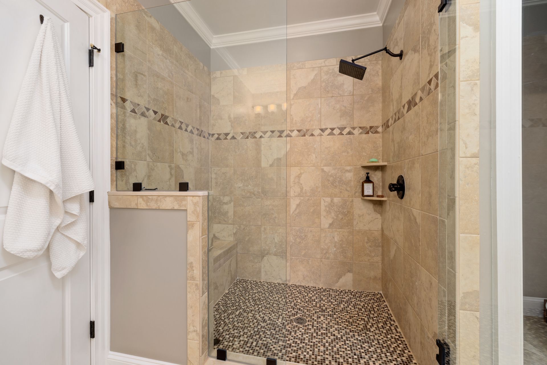 Walk-in shower in beige travertine-look porcelain tile with a diagonal border of brown and cream stone triangles, a tiled half wall and corner bench, a brown glass mosaic floor with a center drain, and frameless glass panels